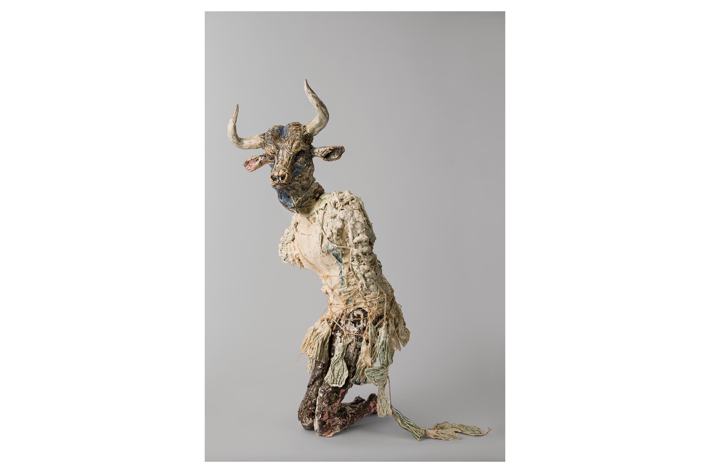A contemporary ceramic sculpture of a kneeling minotaur with a detailed bull’s head and a rough, heavily textured human torso bound in twine against a plain gray background.