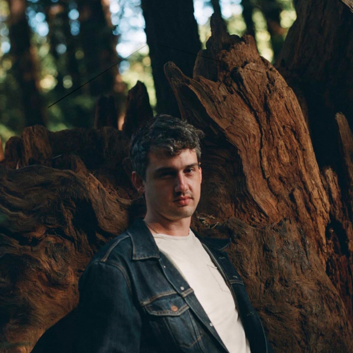 A white man wearing a denim jacket and a white T-shirt looks directly into the camera with a neutral expression. His body is leaning on a large trunk tree.