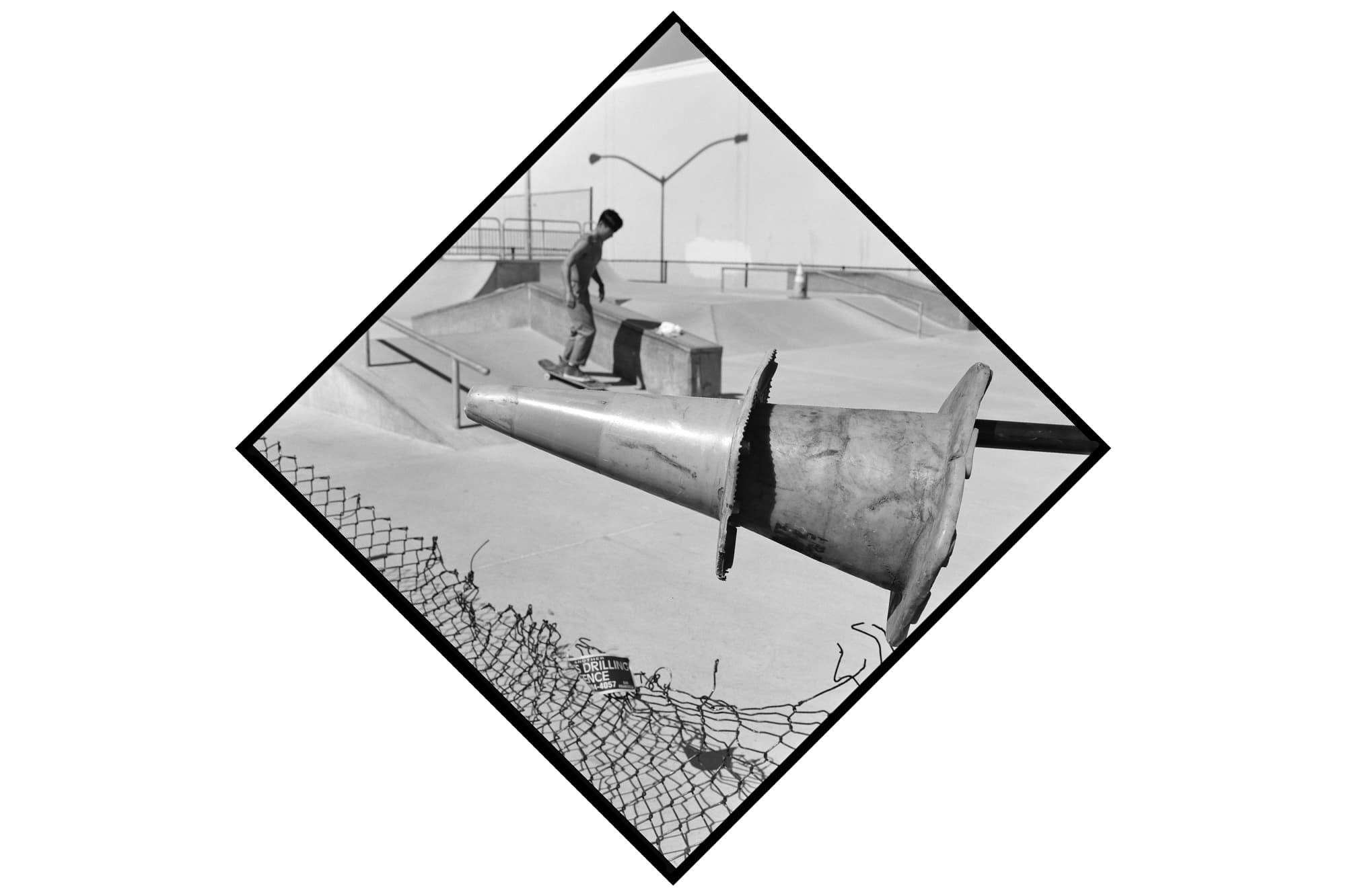A black-and-white photograph of two stacked traffic cones supported by a horizontal pole in the foreground of a skatepark, extending over the edge of a chain-link fence. In the background, a shirtless person rides a skateboard along a concrete ramp, with other ramps and the shadow of a two-armed streetlight visible in the distance.