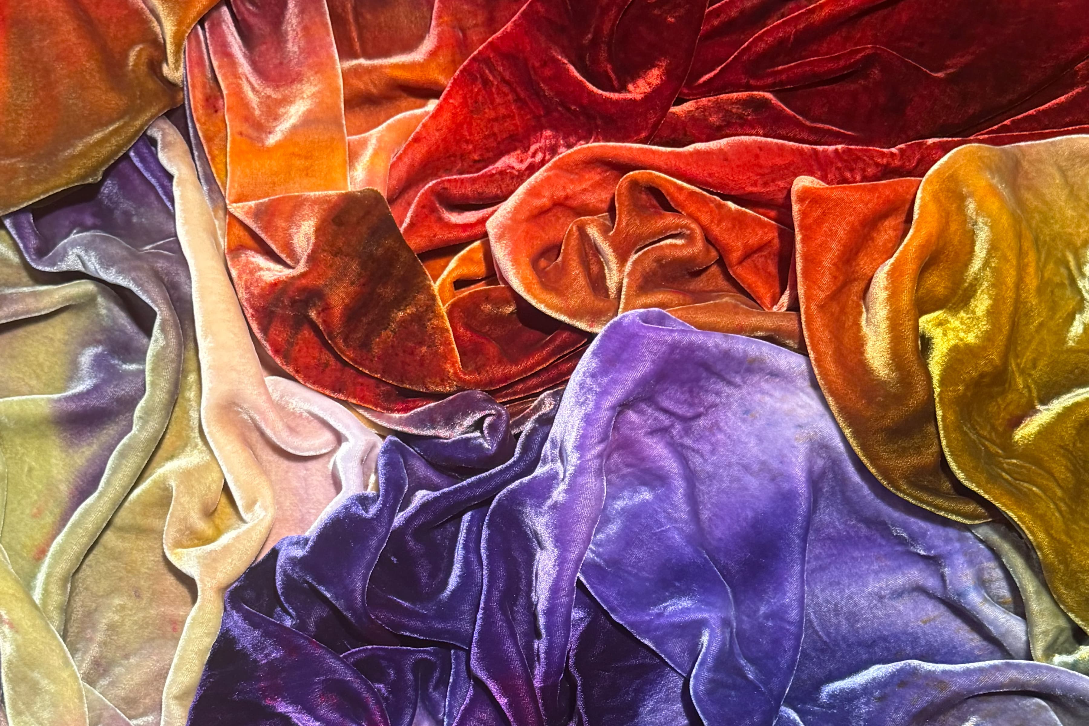 A close-up view of dyed draped silk velvet fabrics in a rich color palette of deep red, burnt orange, golden yellow, plum, and lavender.