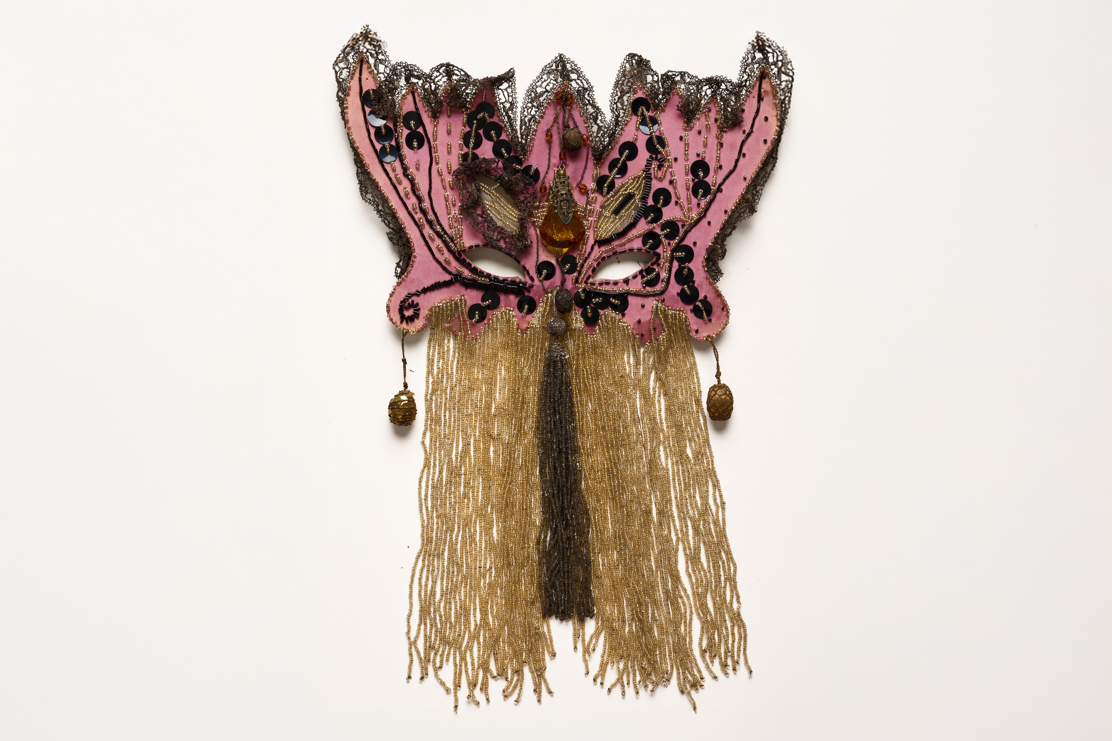 An ornate, butterfly-shaped masquerade mask sits isolated against a solid white background. The mask is primarily crafted from a dusty rose pink fabric, outlined with dark, intricate metallic lace trim that forms pointed crests along the top edge. Black sequins are stitched in swirling patterns across the face of the mask, framing the two eye cutouts. A large, amber-colored gemstone sits directly between the eyes, accented by smaller dark beads. Suspended from the bottom of the mask is a dense fringe of long, metallic gold beads that hangs straight down, creating a shimmering veil. A single, dark brown fabric tassel hangs down the center of the gold fringe, and two small, woven gold beaded ornaments dangle from the lower corners of the mask's wings.