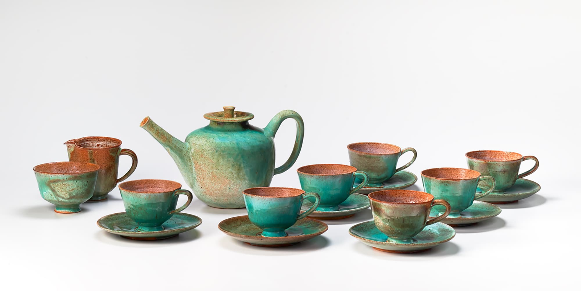 A tea set is arranged on a white background. Central lidded teapot is surrounded by seven matching teacups and saucers, a creamer, and a sugar bowl, with reddish brown glaze on the inside and mottled turquoise around the outside. 