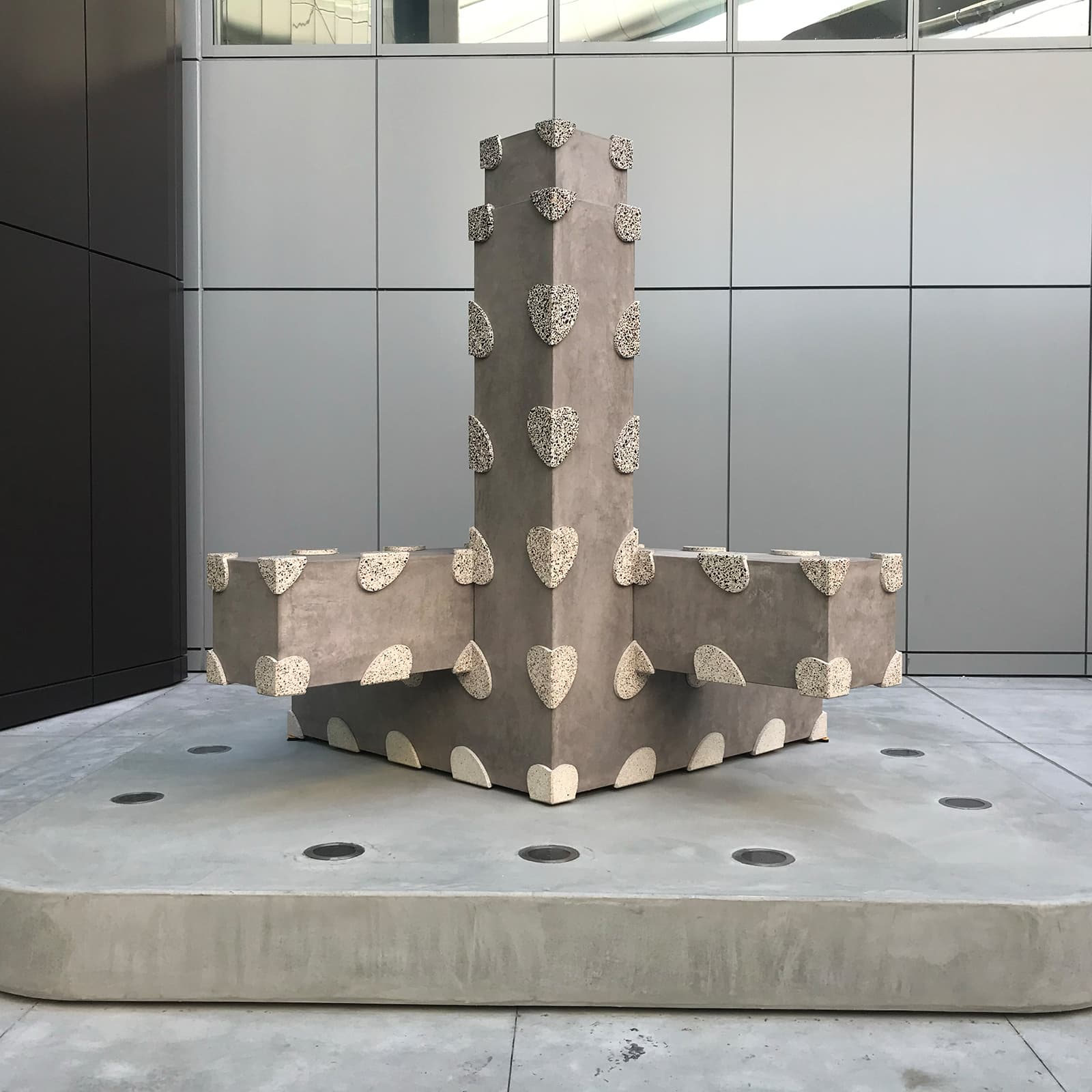 Artwork by Gay Outlaw, A large, gray geometric sculpture shaped like a T-junction or cross sits on an outdoor concrete platform.