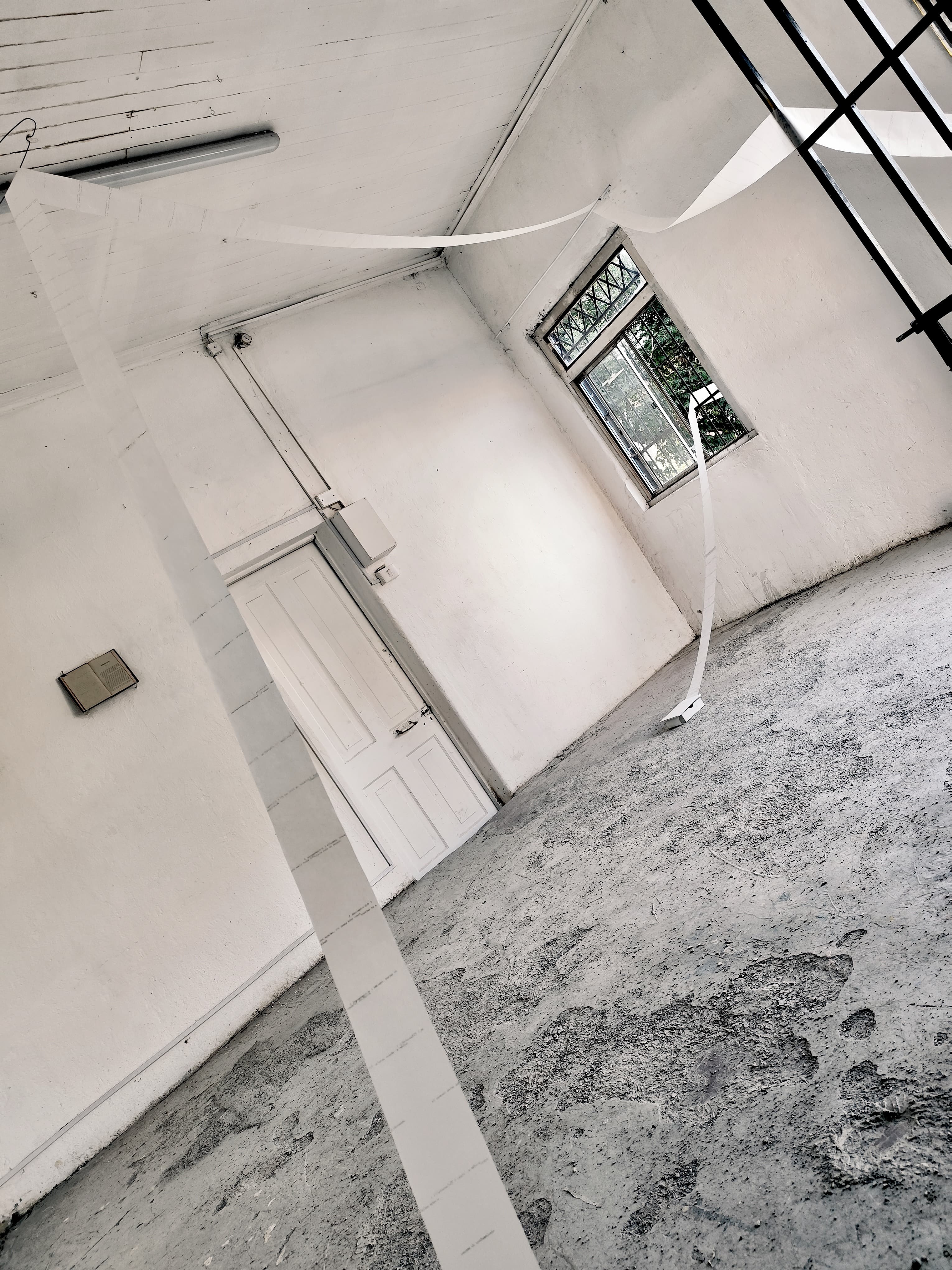 An image of a mostly empty white-walled room, with a narrow sheet of paper coming from a box on the floor, out the window, and returning draped across the space. Faint text is visible on the paper roll.