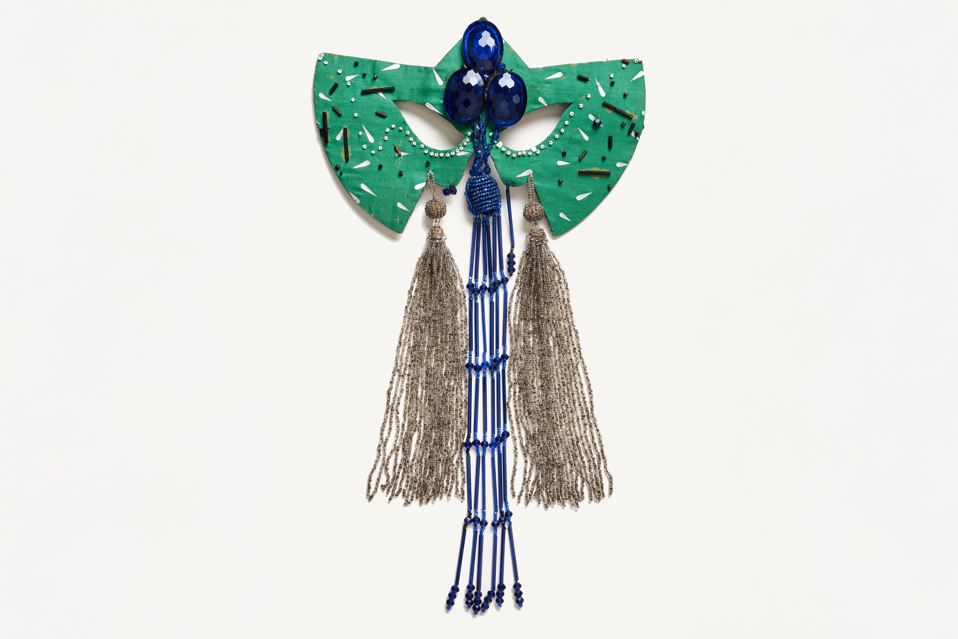 A symmetrical mask shaped out of bright green textured material, resembling abstract wings or a structured bow, embellished with scattered tiny white dots and metallic stick-like beads. Placed directly over the center brow are three large, faceted dark blue teardrop gemstones arranged in a cluster. Cascading vertically from the center of the mask is a long, multi-strand fringe made of tiny blue glass beads, flanked on each side by a dense, elongated tassel of shimmering silver seed beads.