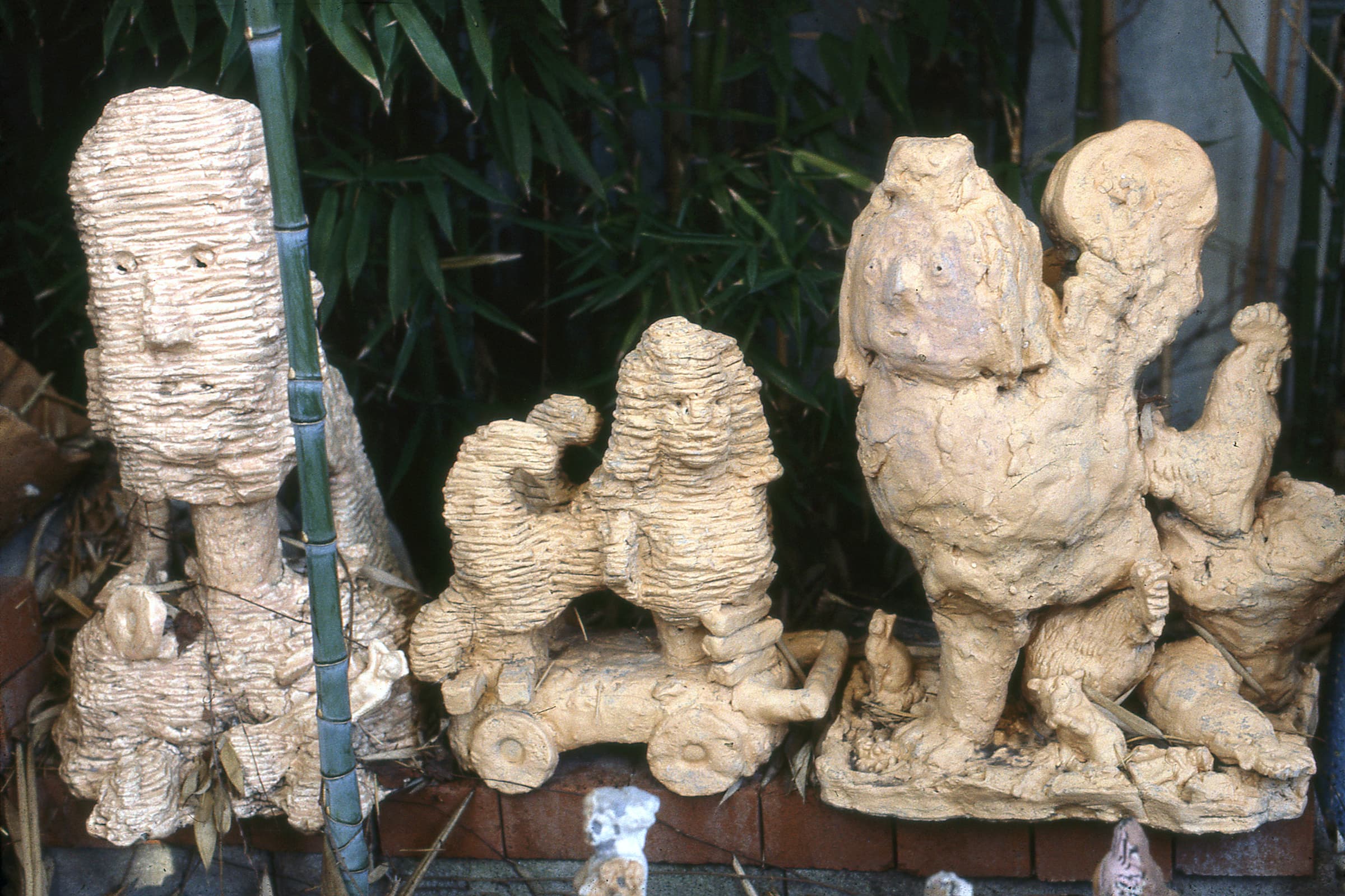 Three light-brown, textured sculptures stand in a row outdoors against a backdrop of green leaves. On the far left, a tall, rectangular head with roughly carved facial features rests on a pedestal, partially framed by a single green bamboo stalk in the foreground. In the center, a stylized animal resembling a poodle or sphinx is built from layered coils of clay and sits atop a wheeled platform. The sculpture on the right features a more abstract, hand-molded figure with smooth and bumpy contours, accompanied by smaller animal-like forms at its base. A low brick border runs along the bottom of the entire display.