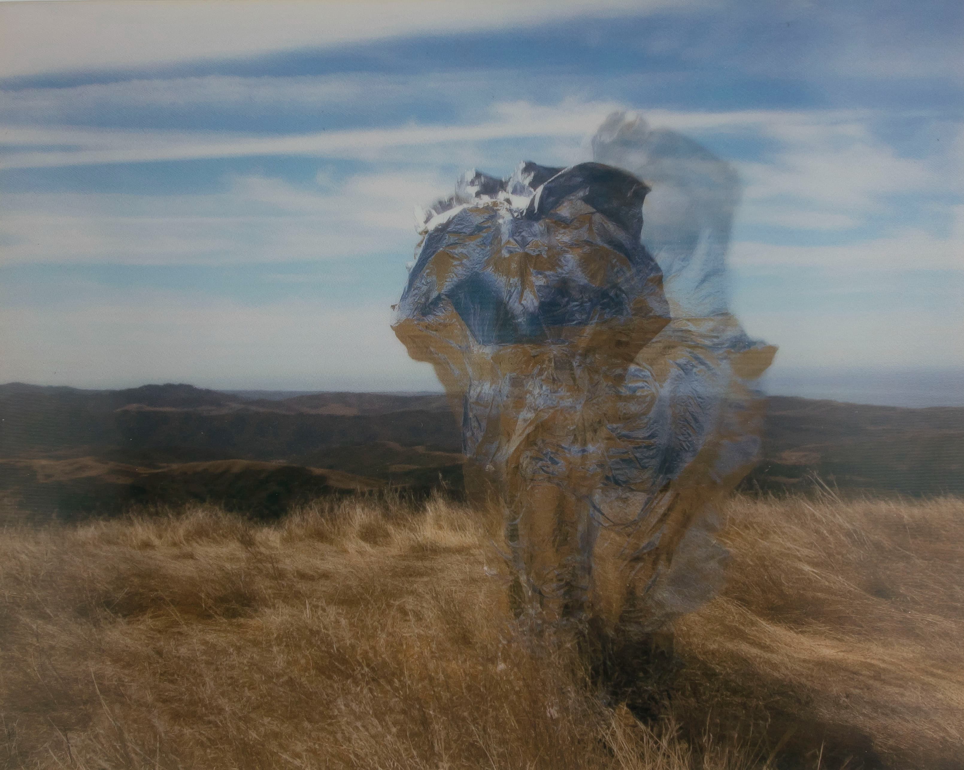 A motion-blurred figure wrapped in reflective silver material moves through a dry, windswept field, with rolling hills and a cloud-streaked blue sky behind them.