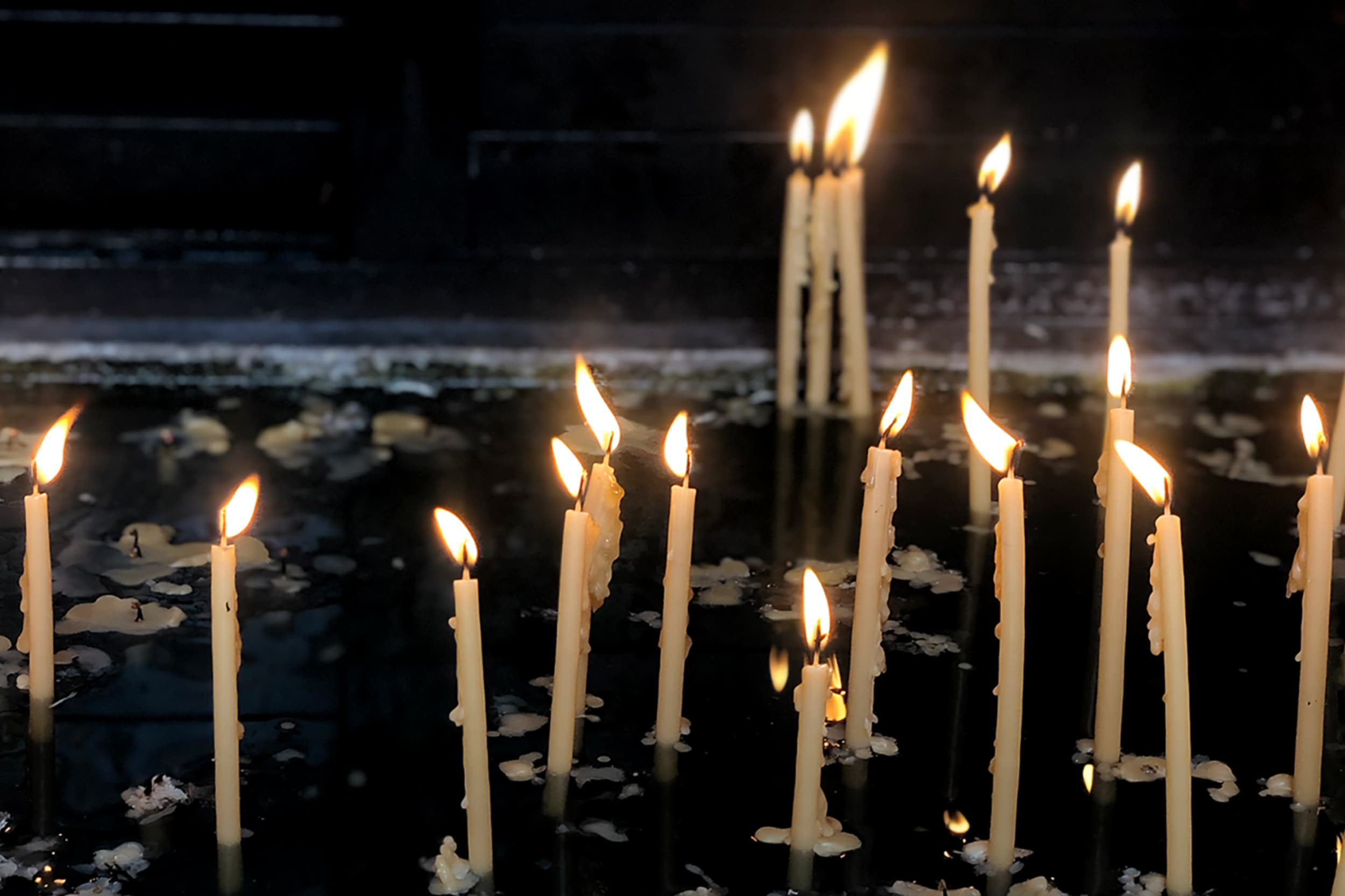 Photograph of long, thin prayer candles burning with wax dripping onto a dark floor.