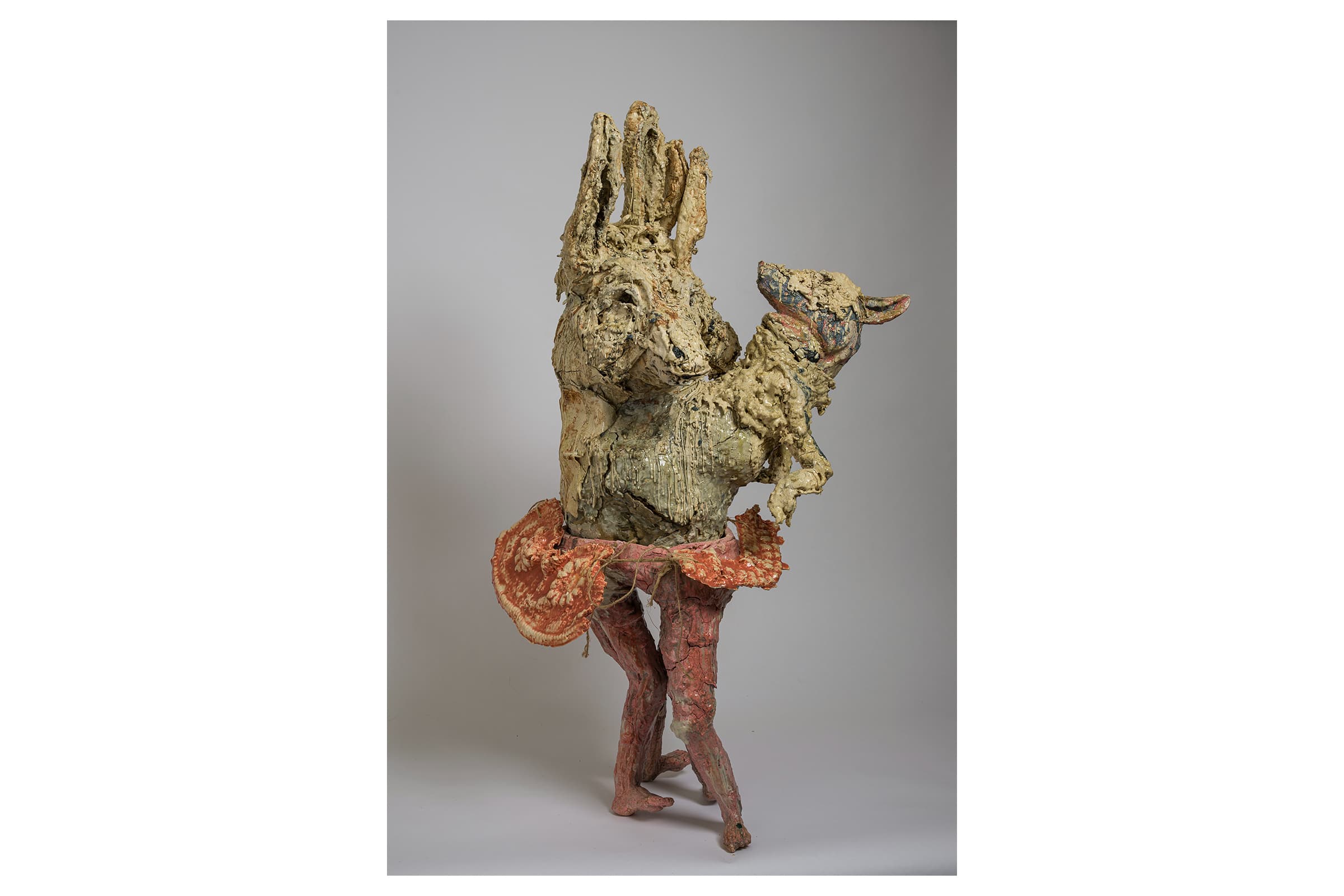 A contemporary mixed-media ceramic sculpture titled "Fugue Dance" by artist Elisabeth Higgins O’Connor, featuring a textured, stylized donkey figure standing on long pinkish legs while wearing a red patterned tutu and holding a small animal figure, isolated against a neutral gray background.