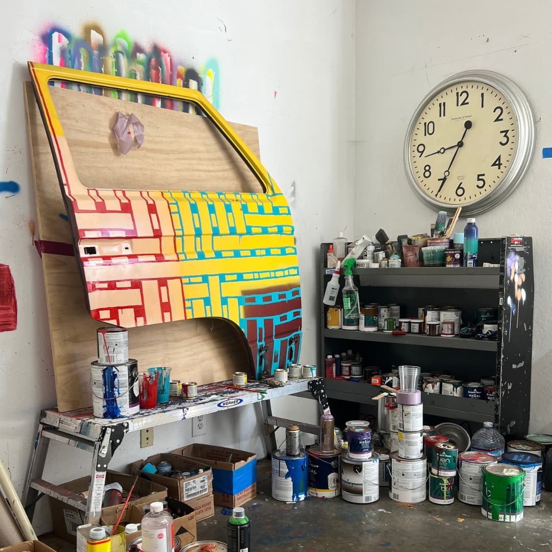 A art studio filled with painting supplies, featuring a large, brightly patchwork painted car door to the left.