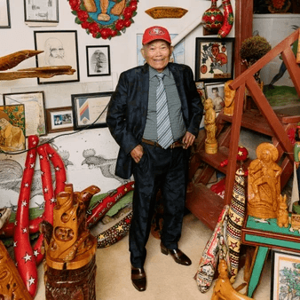 Artist Demitrio Braceros stands in a domestic space surrounded by sculpture and framed works. He wears a suit and a hat.