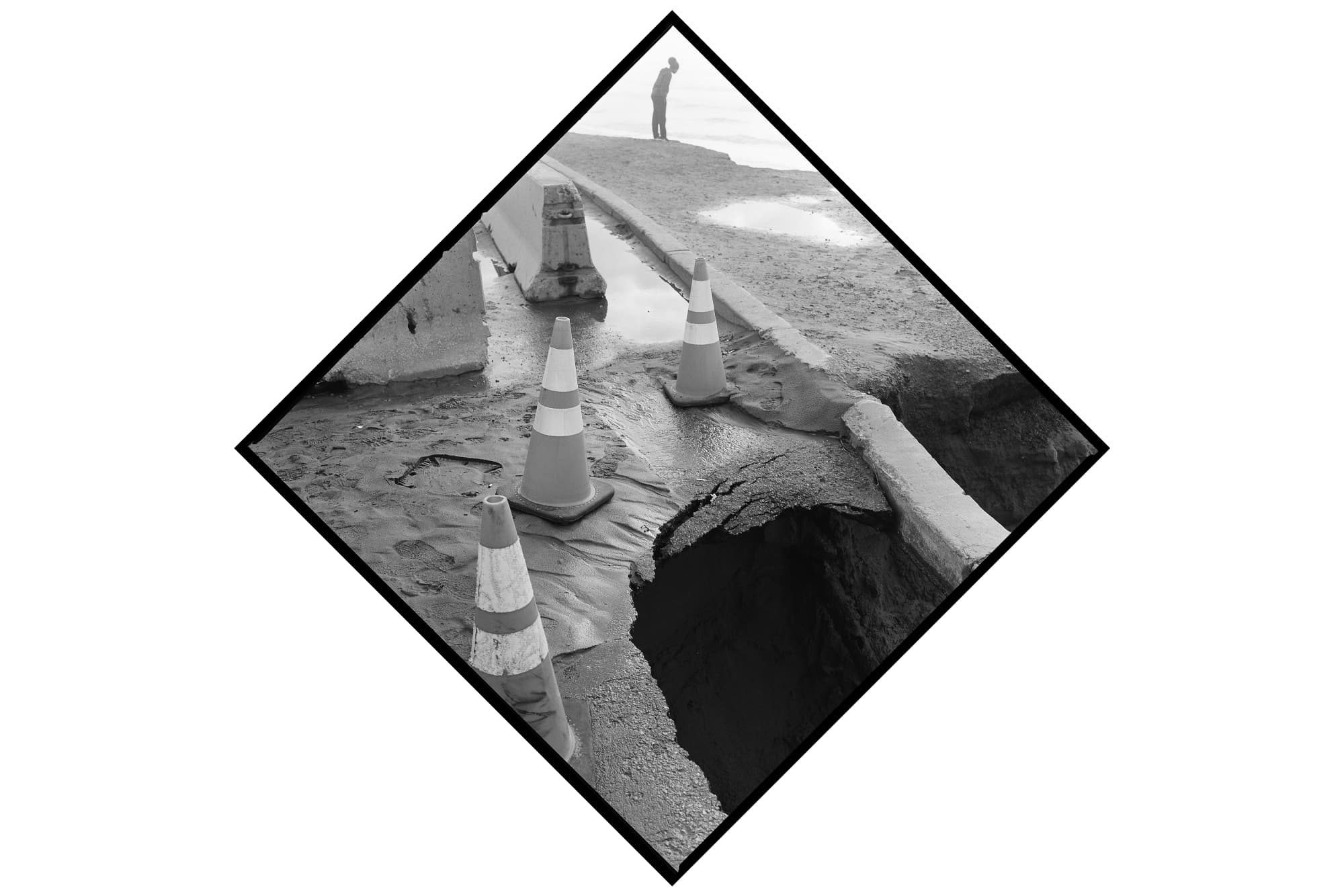 A black-and-white photograph of three traffic cones demarcating the edge of a crumbling sinkhole. In the distance, a partial view of a person appears at the edge of the sand, overlooking the water.