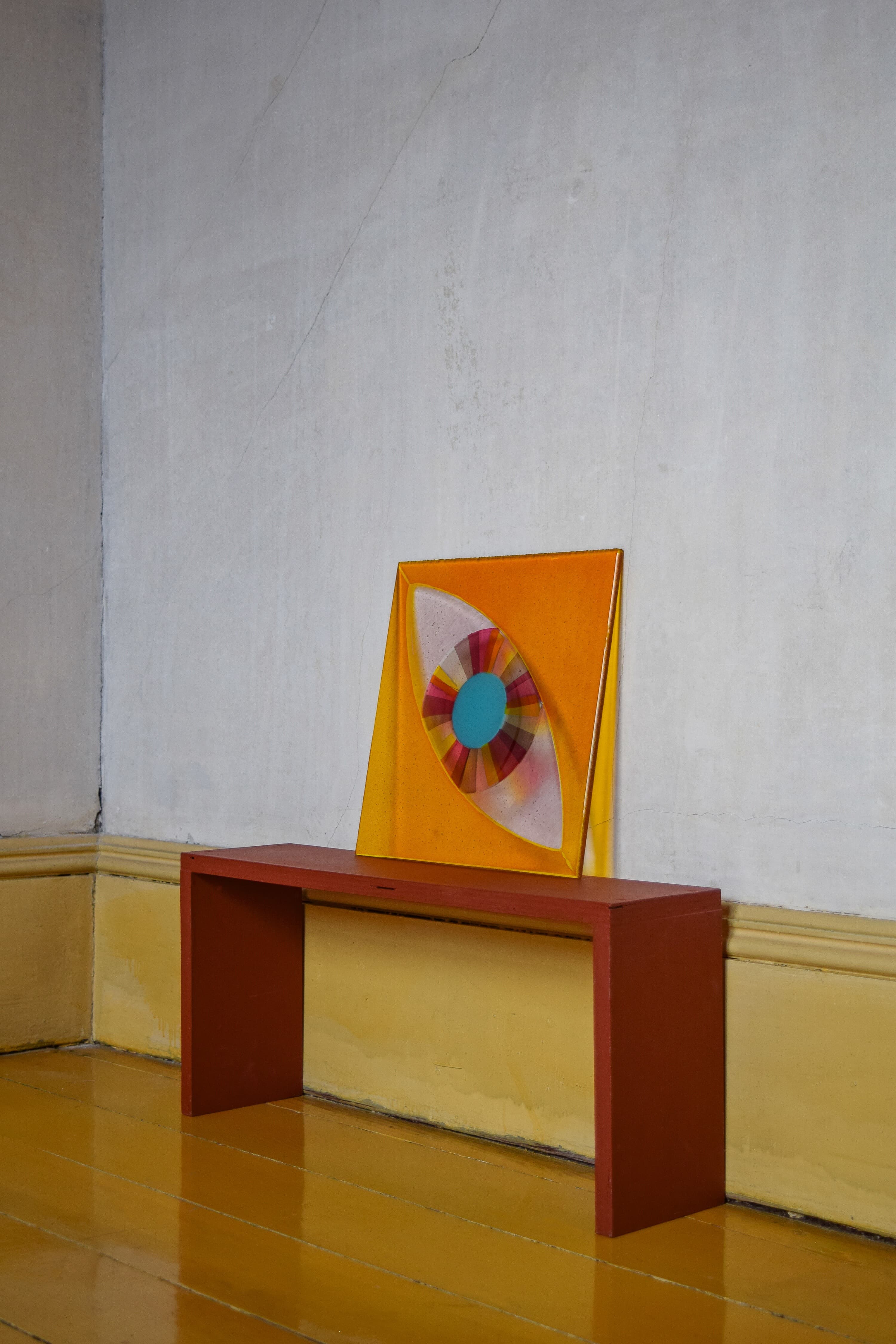 A square orange glass panel with an eye-shaped center, pink and yellow striped iris surround a solid turquoise circle, resting against a wall on a red bench.