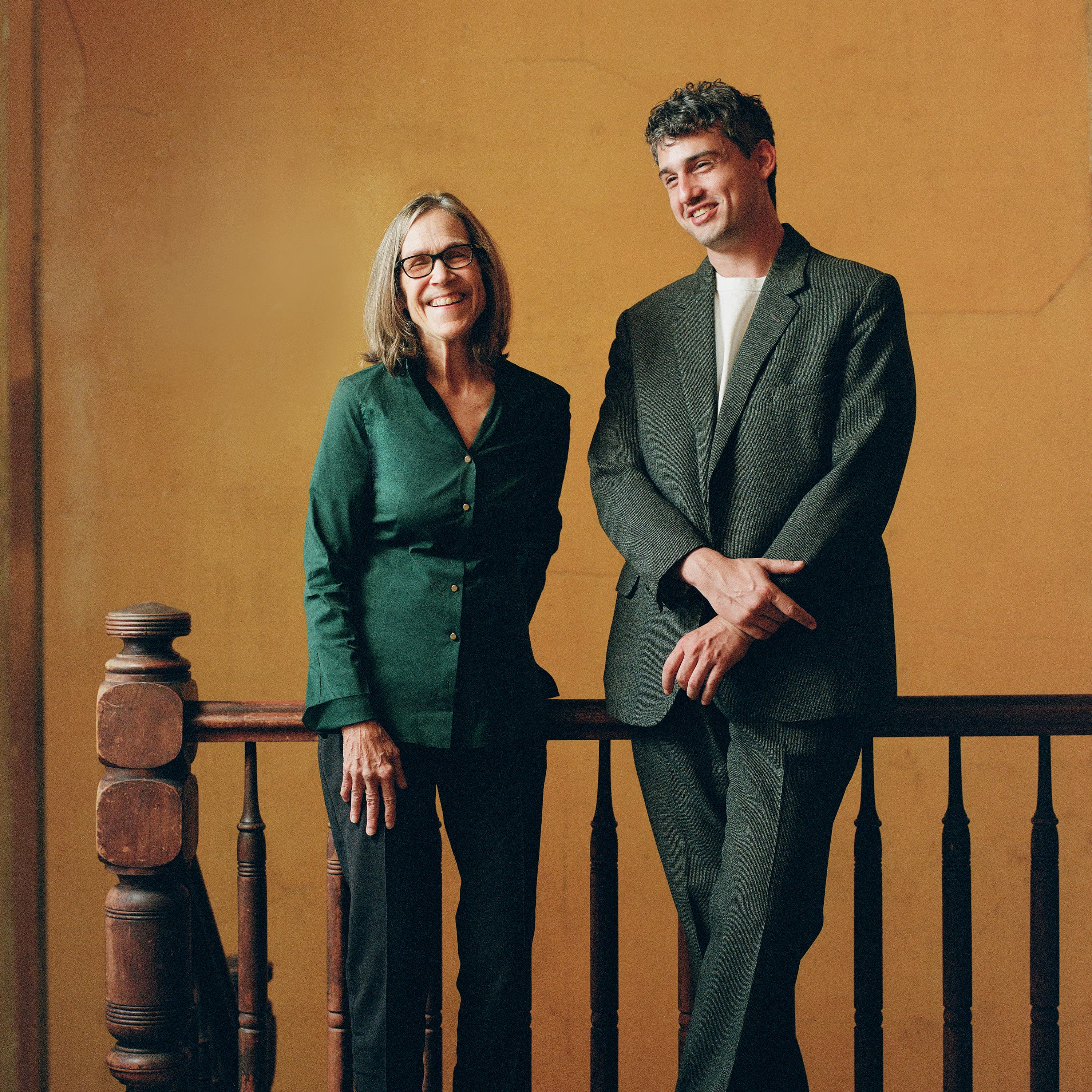 Robin Wright, a middle-aged woman with glasses, and Zully Adler, a man in a grey suit stand next to each other on a balcony in front of a yellow wall. The woman is wearing a dark green shirt and dark pants, and she is smiling. The man is wearing a white undershirt and looking down and to the left with a slight smile. The background is a solid yellow color, and the balcony railing is made of dark wood. The lighting is warm and natural, casting soft shadows on the wall behind them.