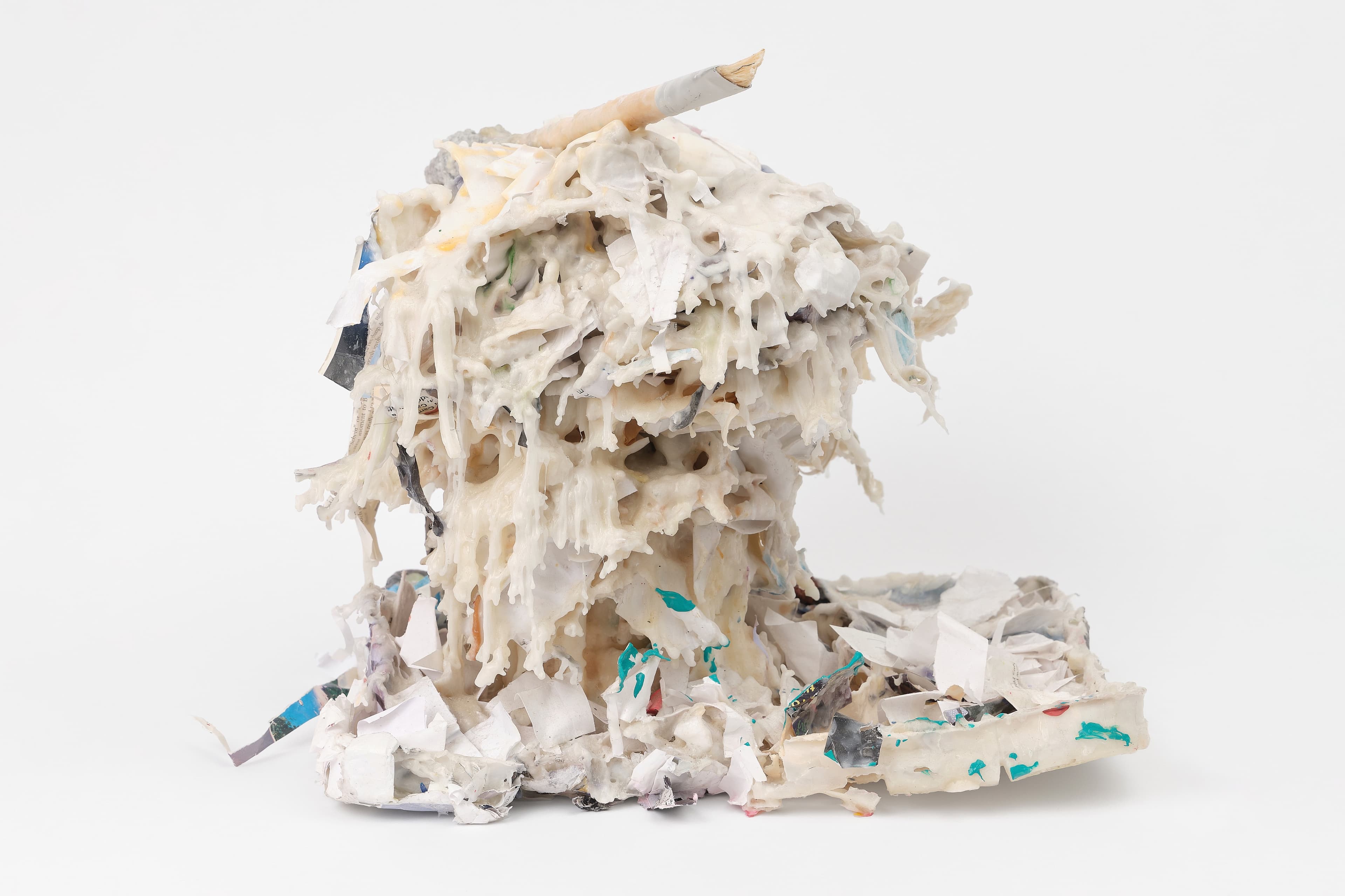 A textured, mushroom-shaped sculpture stands against a plain white background, constructed from a dense accumulation of shredded paper, drips of thick white substance, and colorful debris. The top features a prominent wooden peg, while the melting, column-like structure settles onto a messy base of torn papers and blue or green paint flecks.