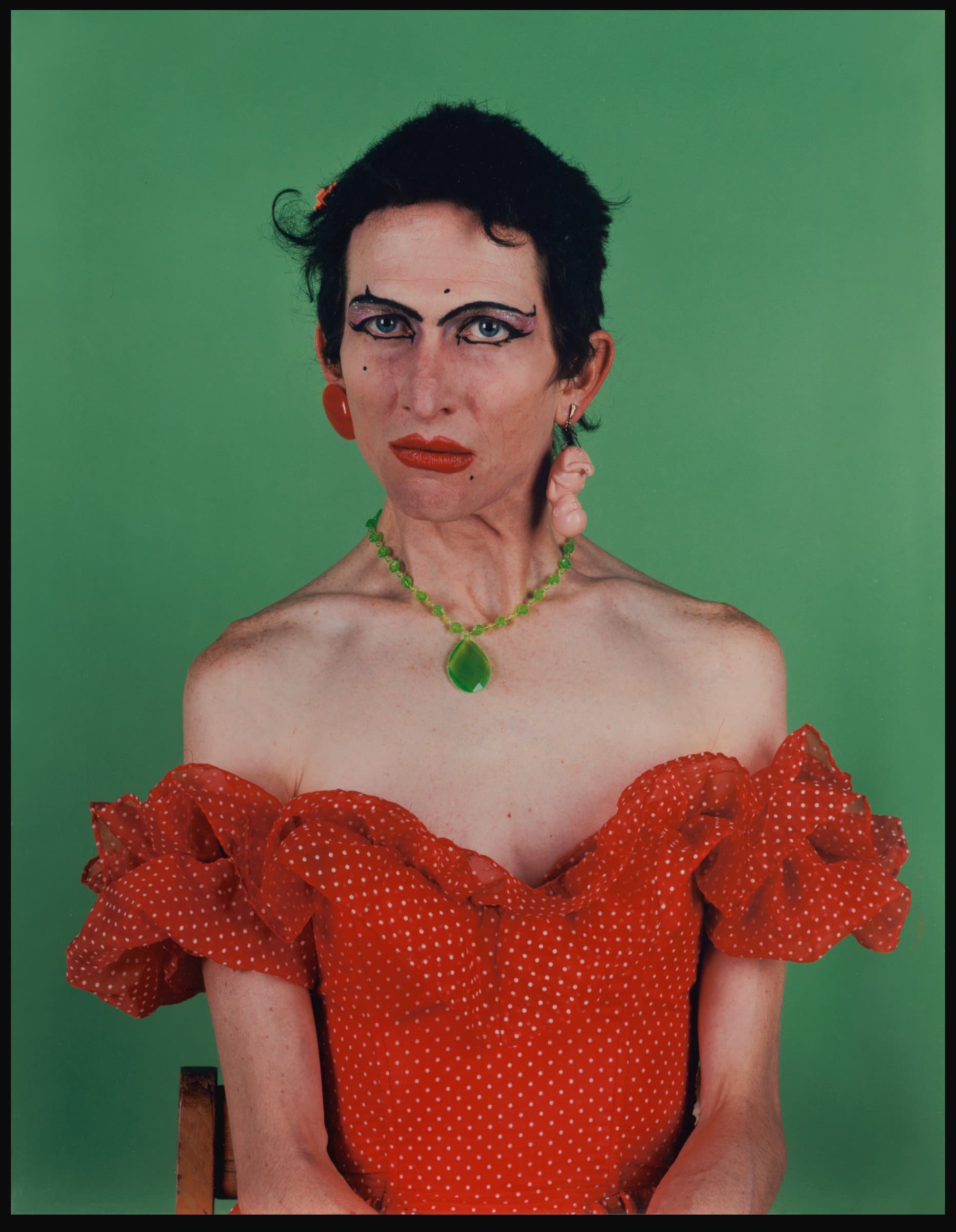 Portrait of a person with short dark hair against a bright green background, wearing dark winged eyeliner, purple eyeshadow, and red lipstick. They wear a red polka-dot off-the-shoulder dress with ruffled sleeves, a beaded green necklace with a teardrop pendant, and mismatched earrings. The subject sits with shoulders bare, gazing directly at the camera.