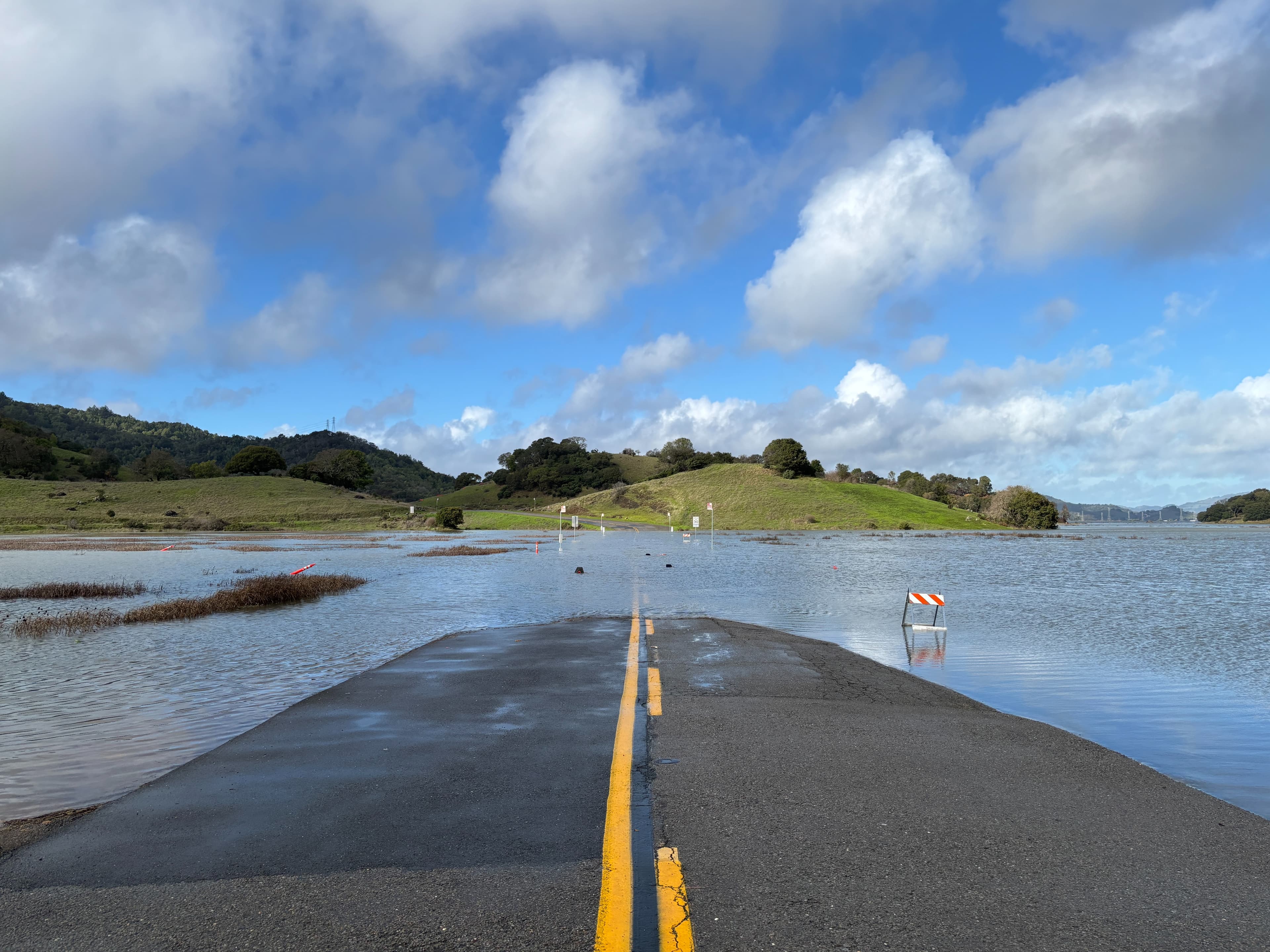 Flooded road (King Tide, China Camp State Park, Jan 3, 2026): A paved road narrows to a point where it disappears entirely under floodwater, its double yellow line running straight toward a submerged warning sign; green hills and oak trees rise beyond the waterline under a partly cloudy sky.