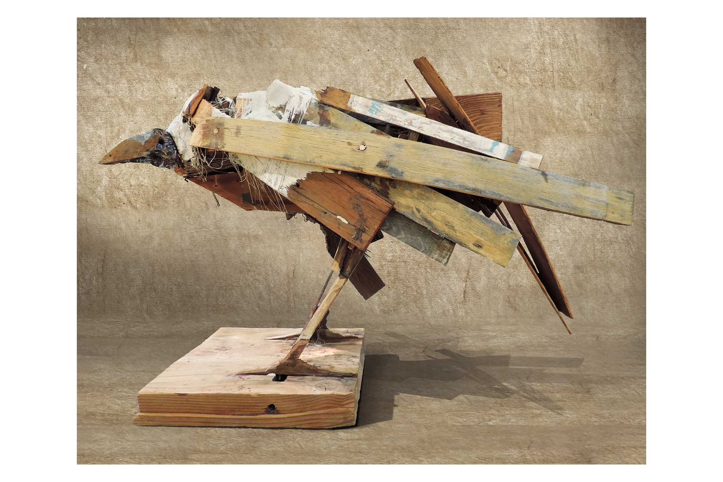 A contemporary assemblage sculpture of a bird made from scrap lumber, plaster, and metal. The abstract bird figure is mounted on a flat wooden base and positioned in profile, facing left against a textured, neutral-toned wall.