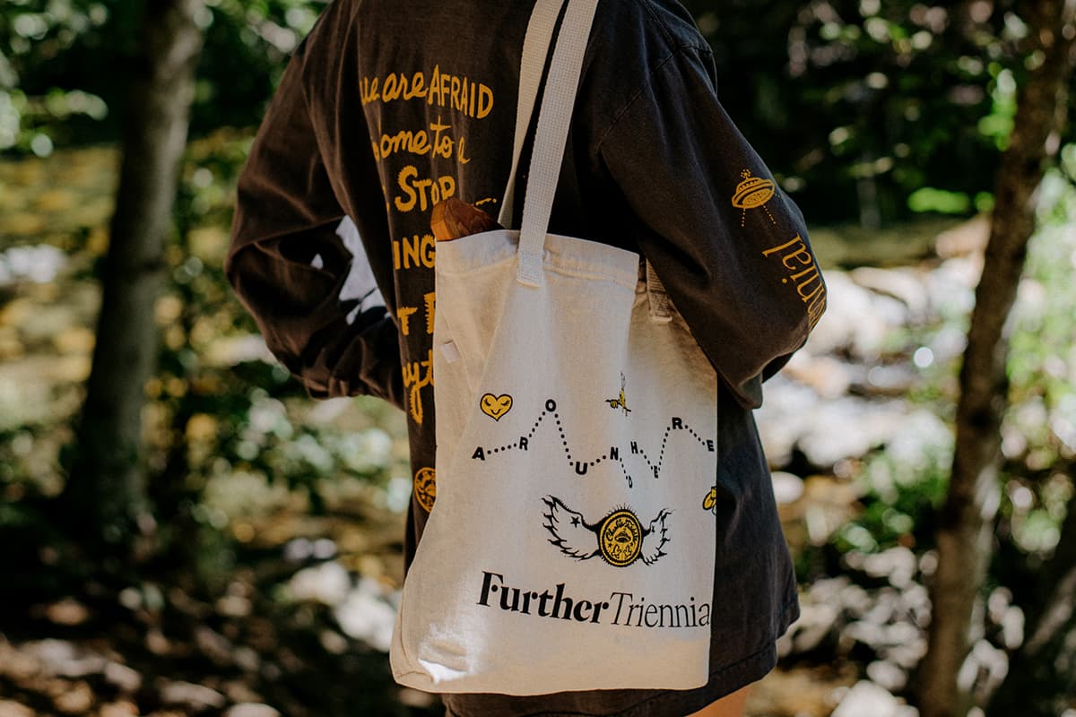 A person giving their back to the camera wears Further branded black shirt and canvas bag. This person is somewhere in a park or forest.