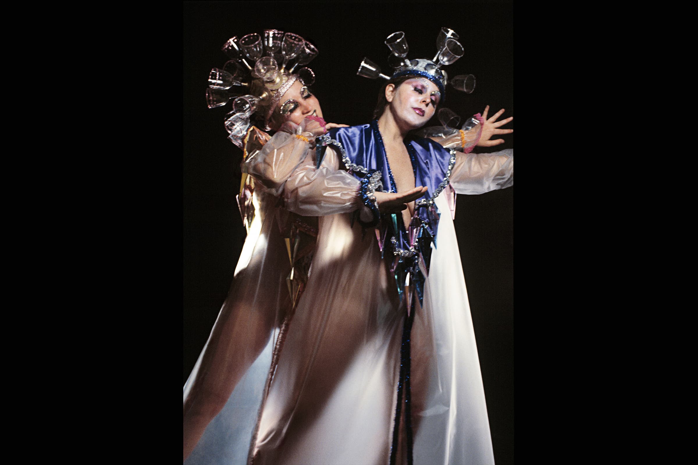 Two performers wearing avant-garde costumes with wine glass headpieces embrace against a solid black background. The performer on the right has pale white facial makeup and dark lipstick, wearing a long white gown adorned with a purple satin collar and a dangling collar of geometric, prismatic metallic spikes. The performer on the left reaches over from behind, clad in a sheer, iridescent overlay. Both wear elaborate, crown-like headpieces branching outwards, completely constructed from upside-down, transparent wine glasses. The lighting is focused heavily on the performers, accentuating the reflective glass elements and the flowing fabric of their gowns.