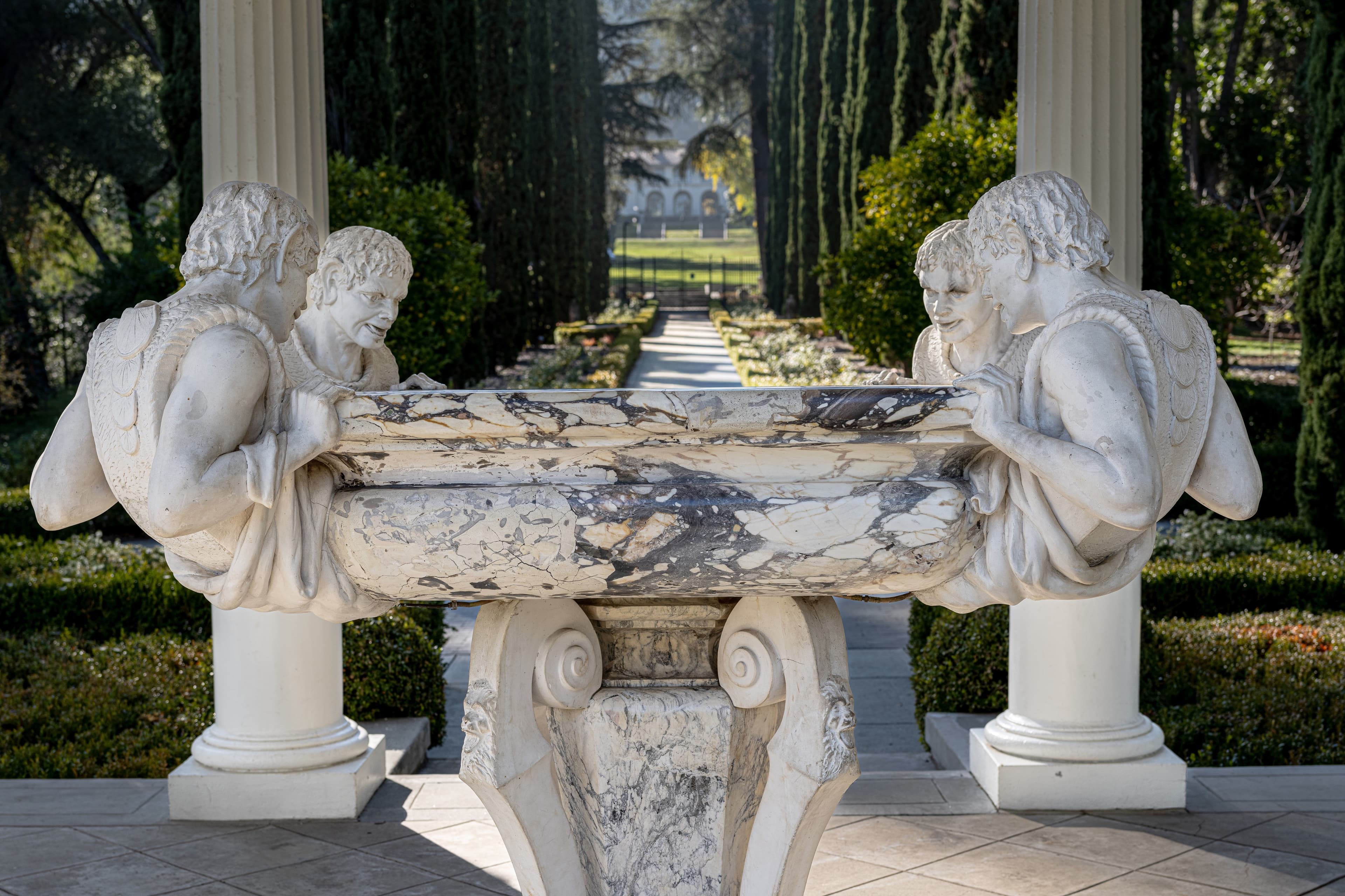 Marble fountain with four fantastical figures hanging on each corner of an oval-shaped basin.