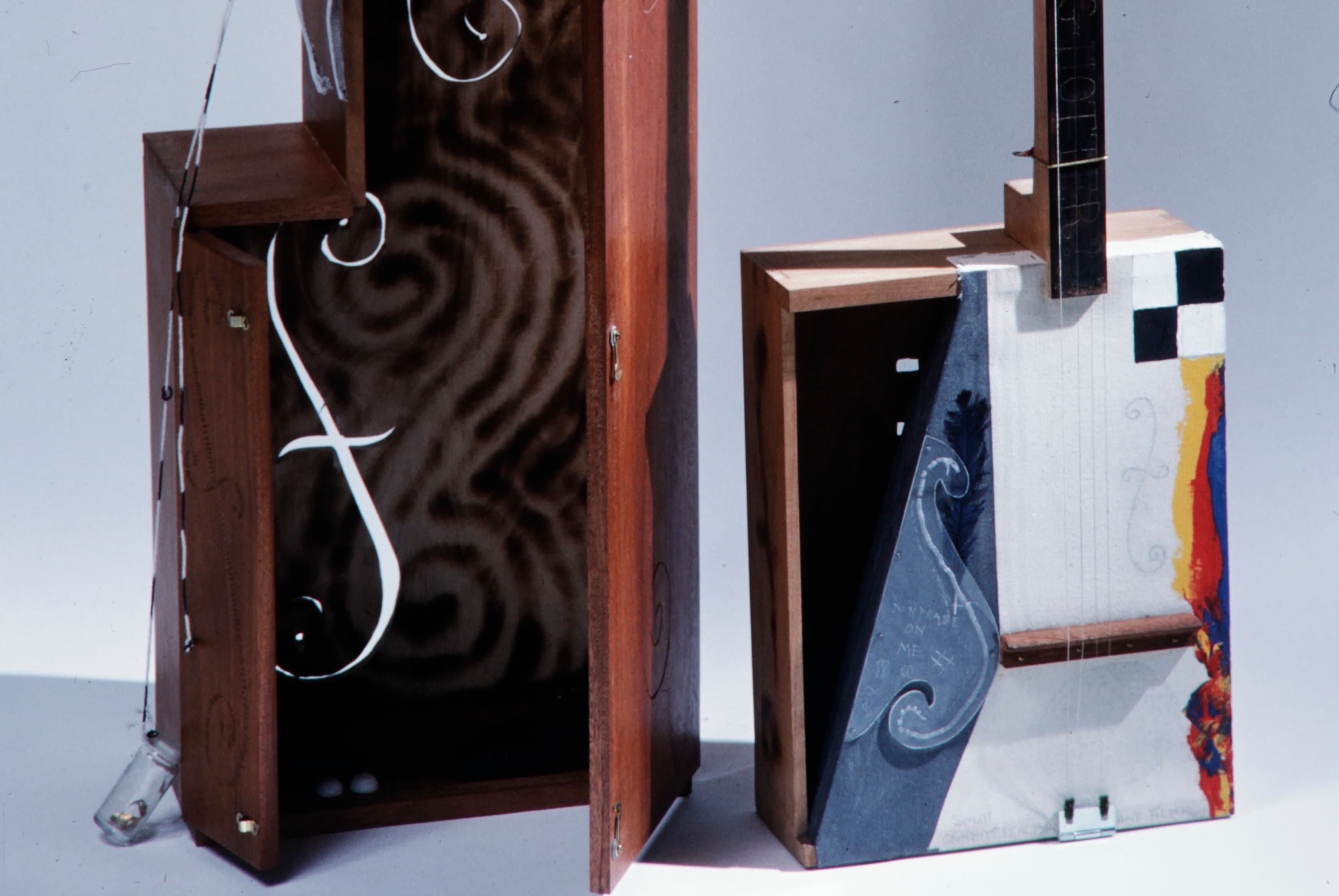 A close-up photograph of two custom wooden stringed instruments with mixed-media art. On the left, an open reddish-brown case reveals a marbled interior and curving white wires with a hanging glass vial. On the right, a rectangular instrument features a white face with strings, a grey section painted with a violin f-hole, a black-and-white checkerboard corner, and a splash of colorful abstract paint.