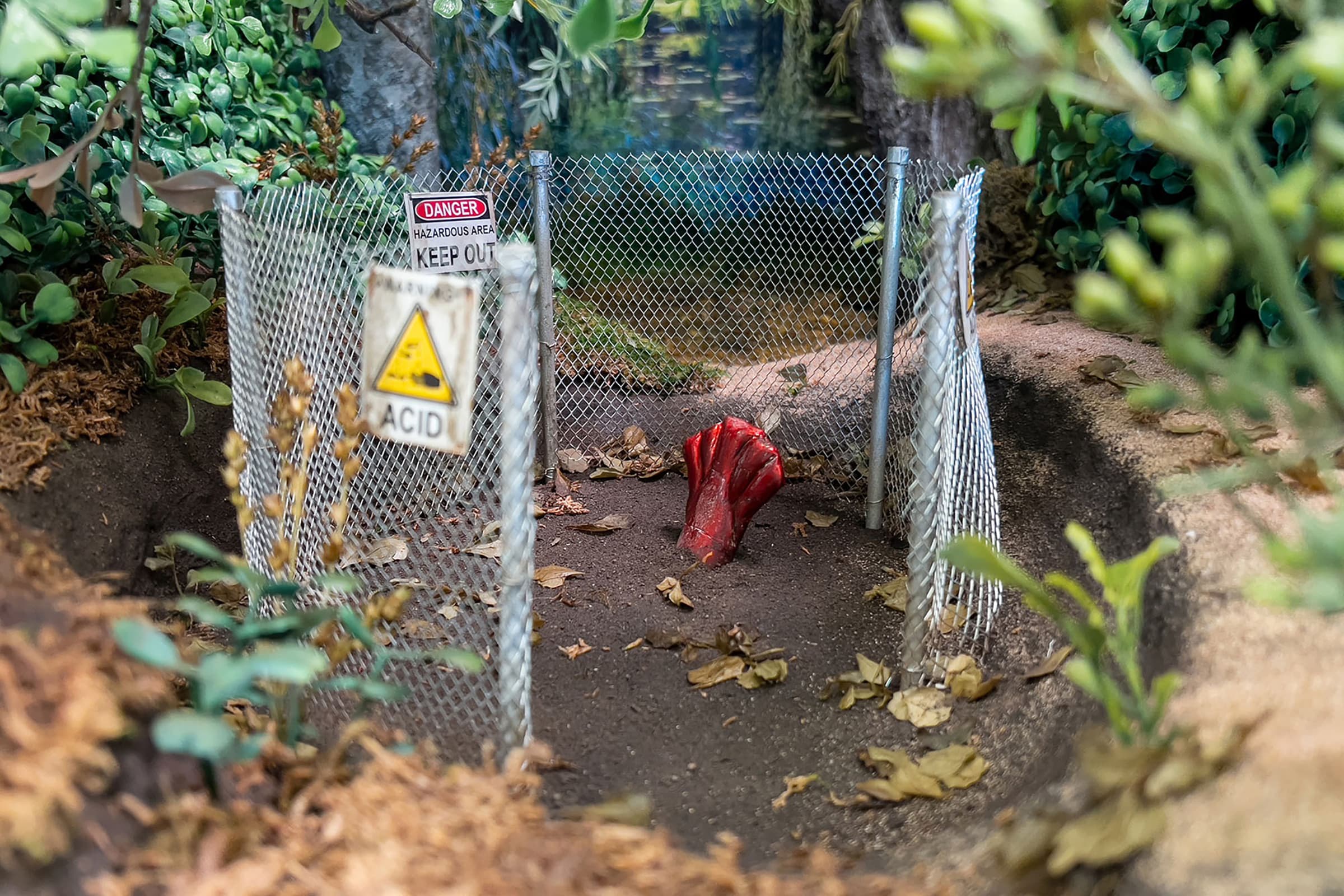 A miniature diorama depicts a small, circular chain-link fence enclosing a patch of dark soil, out of which a glossy red, claw-like structure emerges. Miniature warning signs are attached to the fence posts, including a red-and-white "DANGER HAZARDOUS AREA KEEP OUT" sign and a yellow triangular "ACID" warning sign. The fenced-in area sits within a depression in the ground, surrounded by realistic faux greenery, moss, and sandy terrain that creates a realistic, scale-model outdoor environment.
