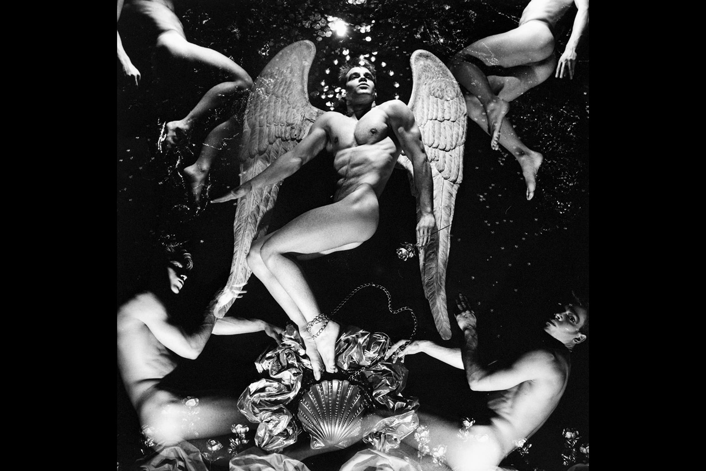 A black and white photograph featuring a muscular, winged male figure resembling an angel lying down at the center, surrounded by four other nude figures in a dynamic, circular composition against a dark backdrop. The central figure has large, textured feathery wings outstretched, a chain wrapped around his ankle, and holds a rose, while the surrounding figures frame him from the corners of the image amidst scattered flowers and a metallic shell shape at the bottom of his feet.