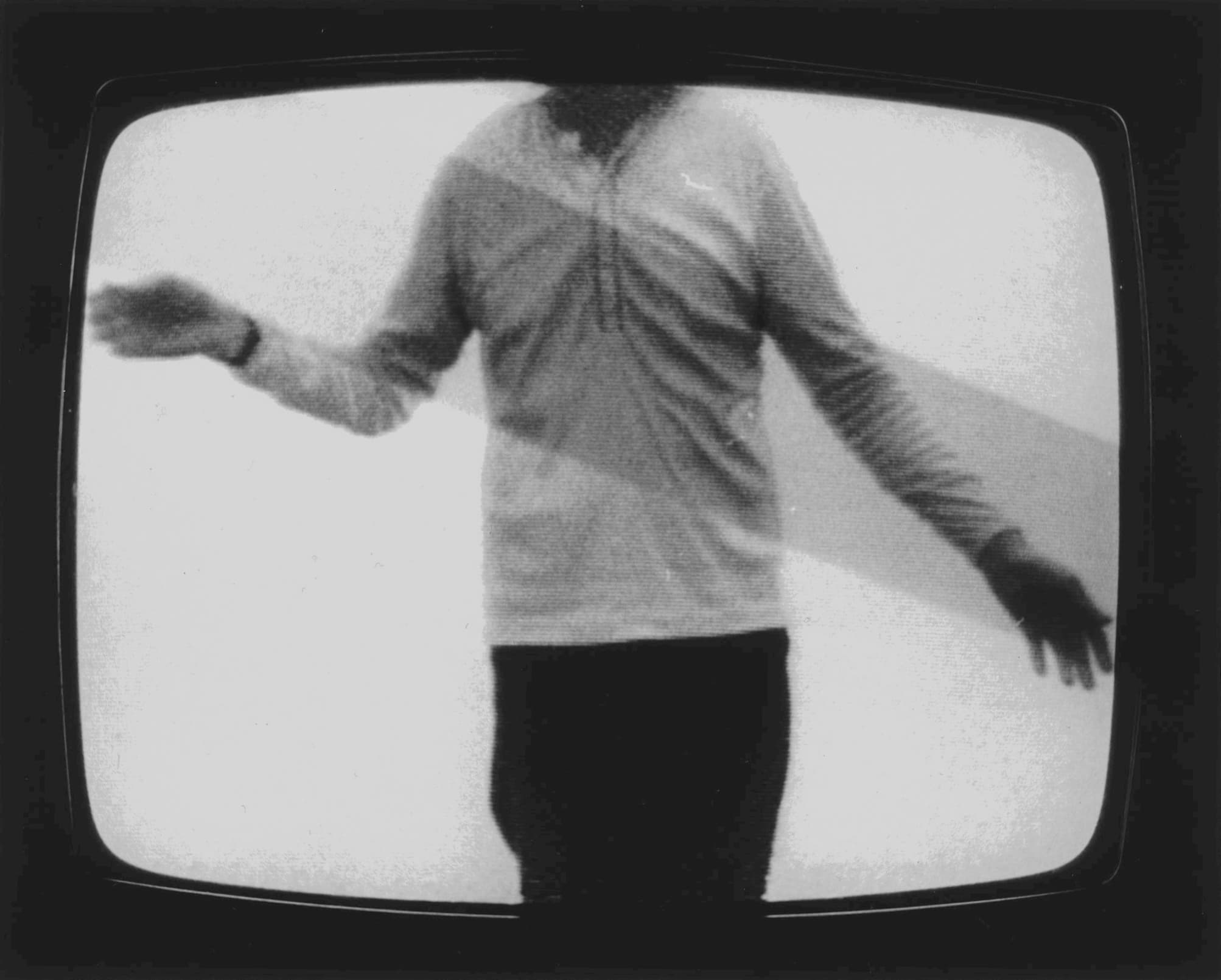 Black-and-white photograph of a video playing on a cube monitor. Film features the torso, arms, and top of legs of a male-presenting figure wearing black pants and long-sleeved henley shirt.