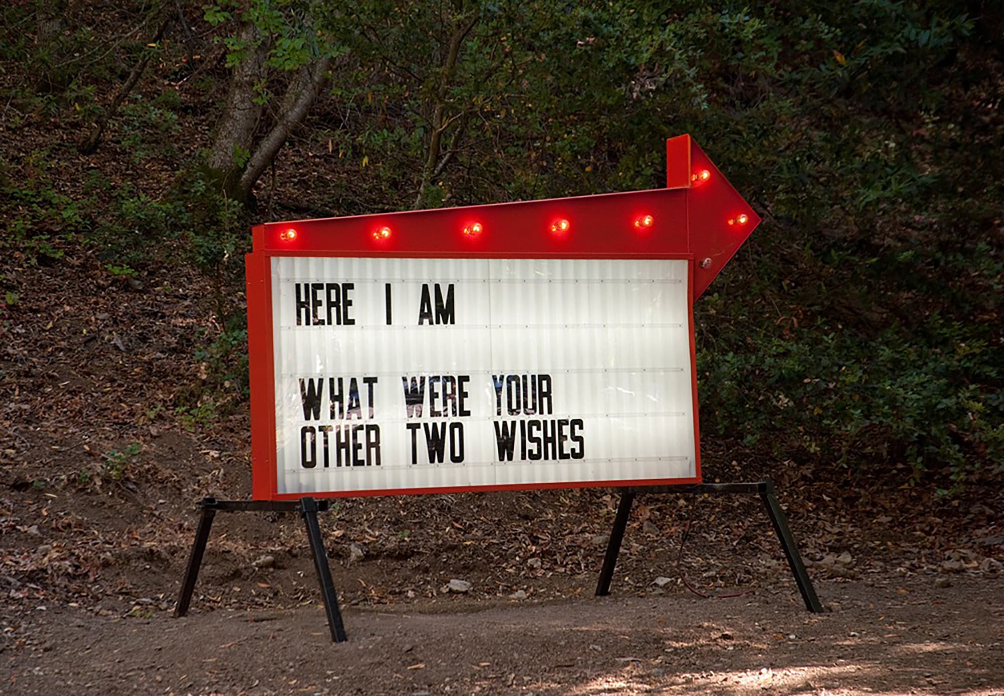 Illuminated directional sign with a red arrow displaying the message, “HERE I AM WHAT WERE YOUR OTHER TWO WISHES?”