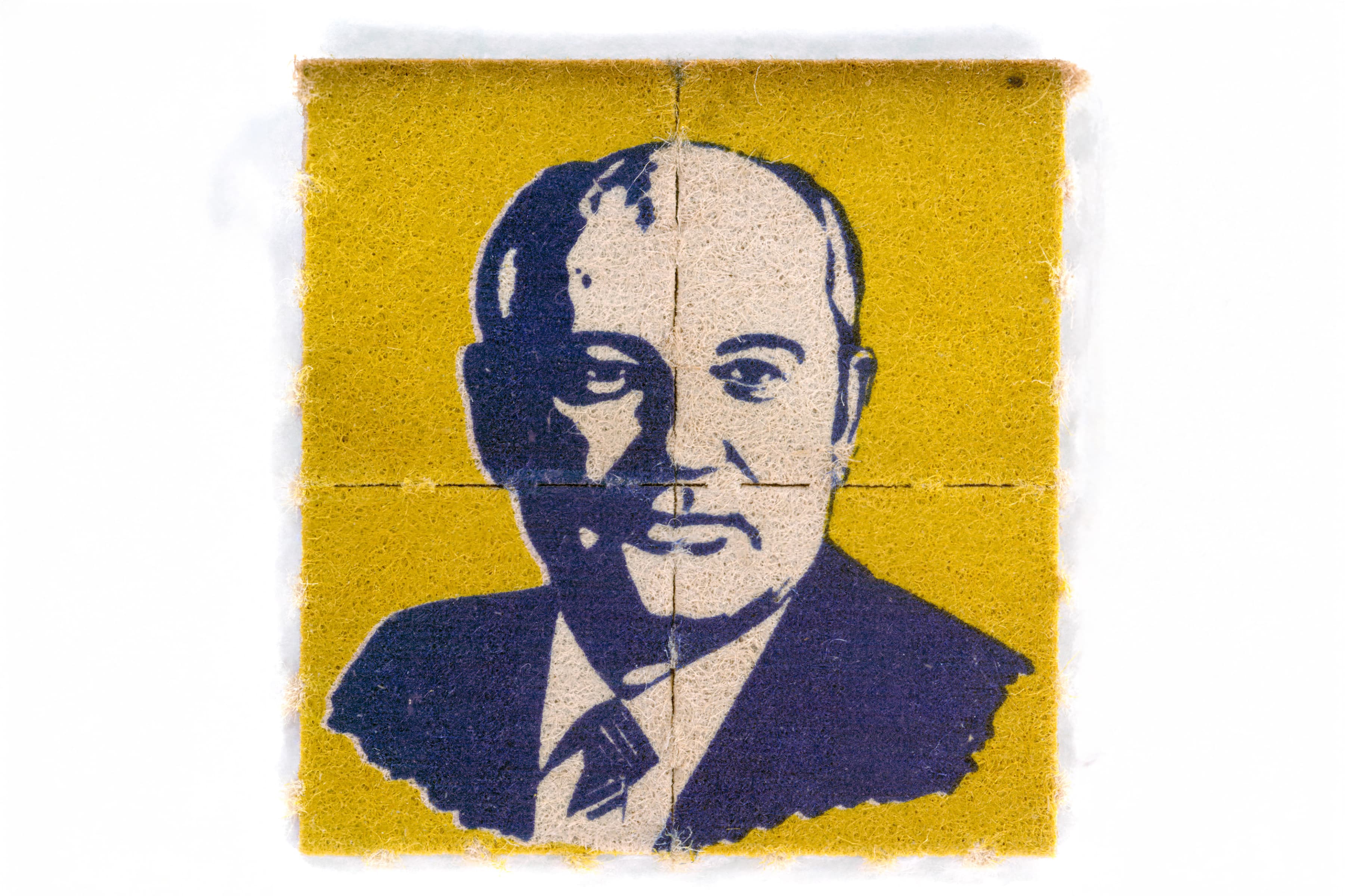 A square piece of yellow blotter paper divided by faint cross-perforations into four smaller squares. A stylized, high-contrast portrait of Mikhail Gorbachev is printed across the sheet in dark blue ink.