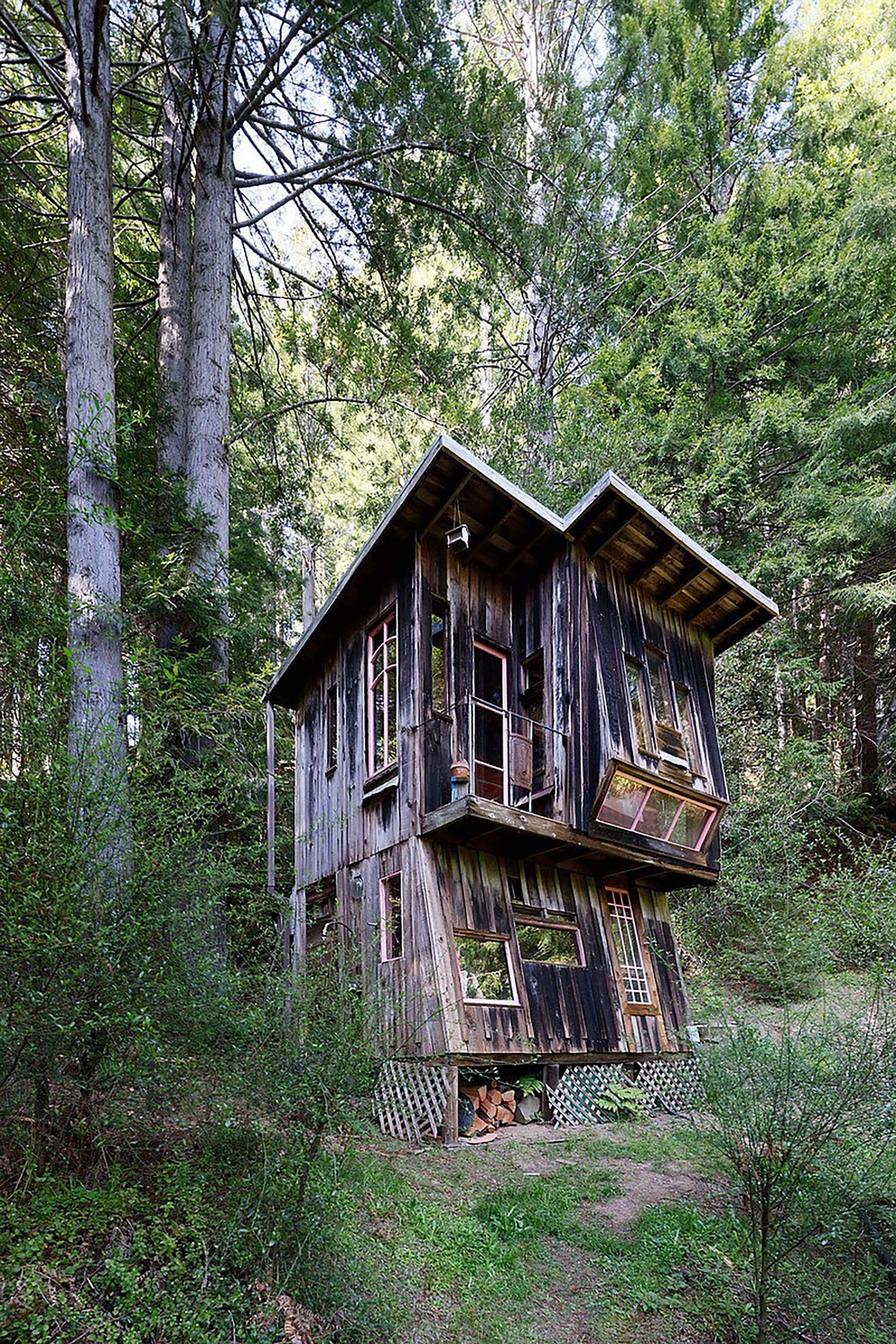 Photograph of a rustic, two-story wooden cabin in a dense forest, featuring an unconventional, angled structure with windows on both levels.