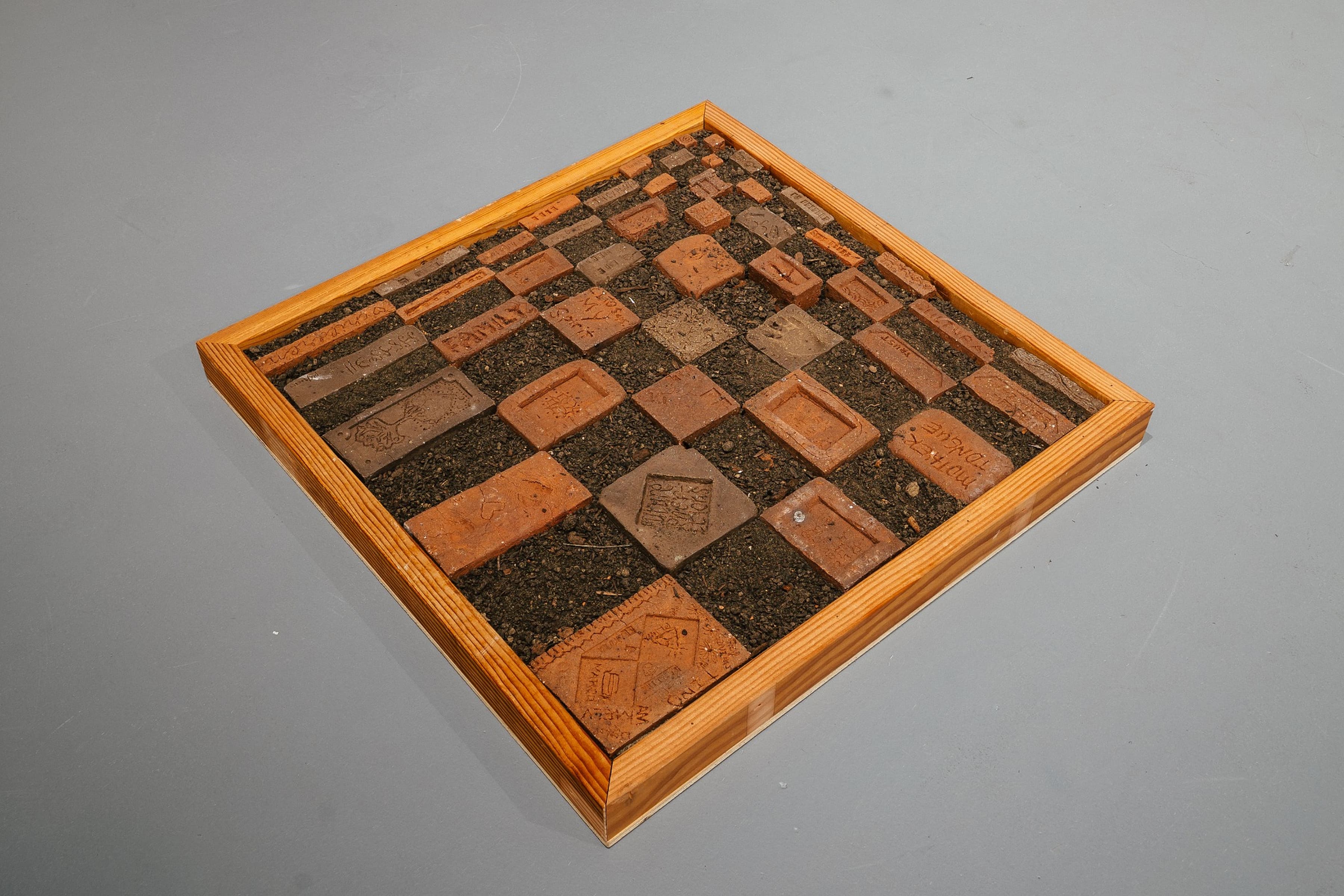 A square wooden frame resembling a chess board sits on a gray floor, filled with soil and brick arrangements of various sizes, some stamped with text like "FAMILY" and "MOTHER TONGUE."