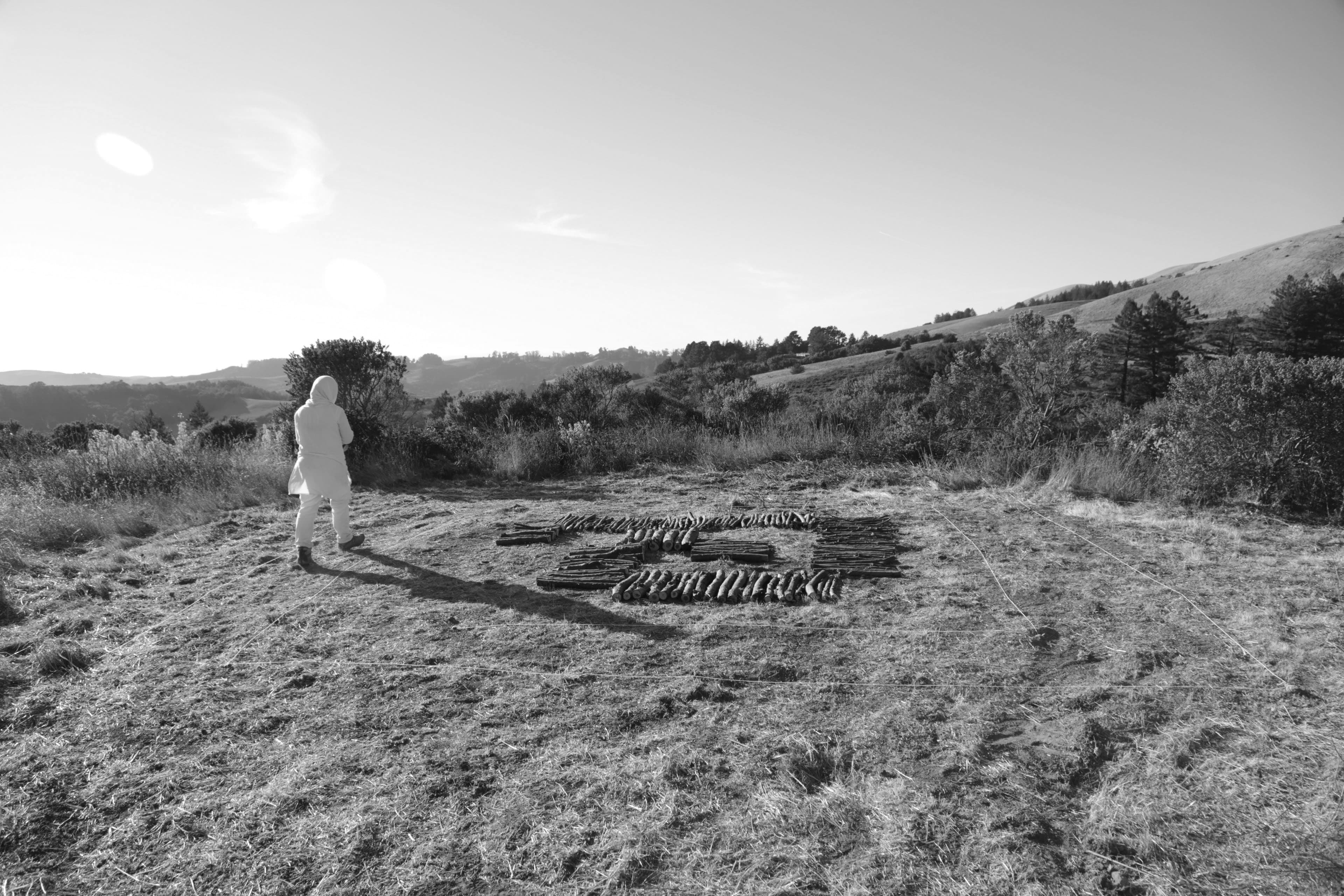 A black-and-white photograph of a hooded figure standing beside logs arranged in a geometric pattern on the ground, surrounded by rolling hills and vegetation.
