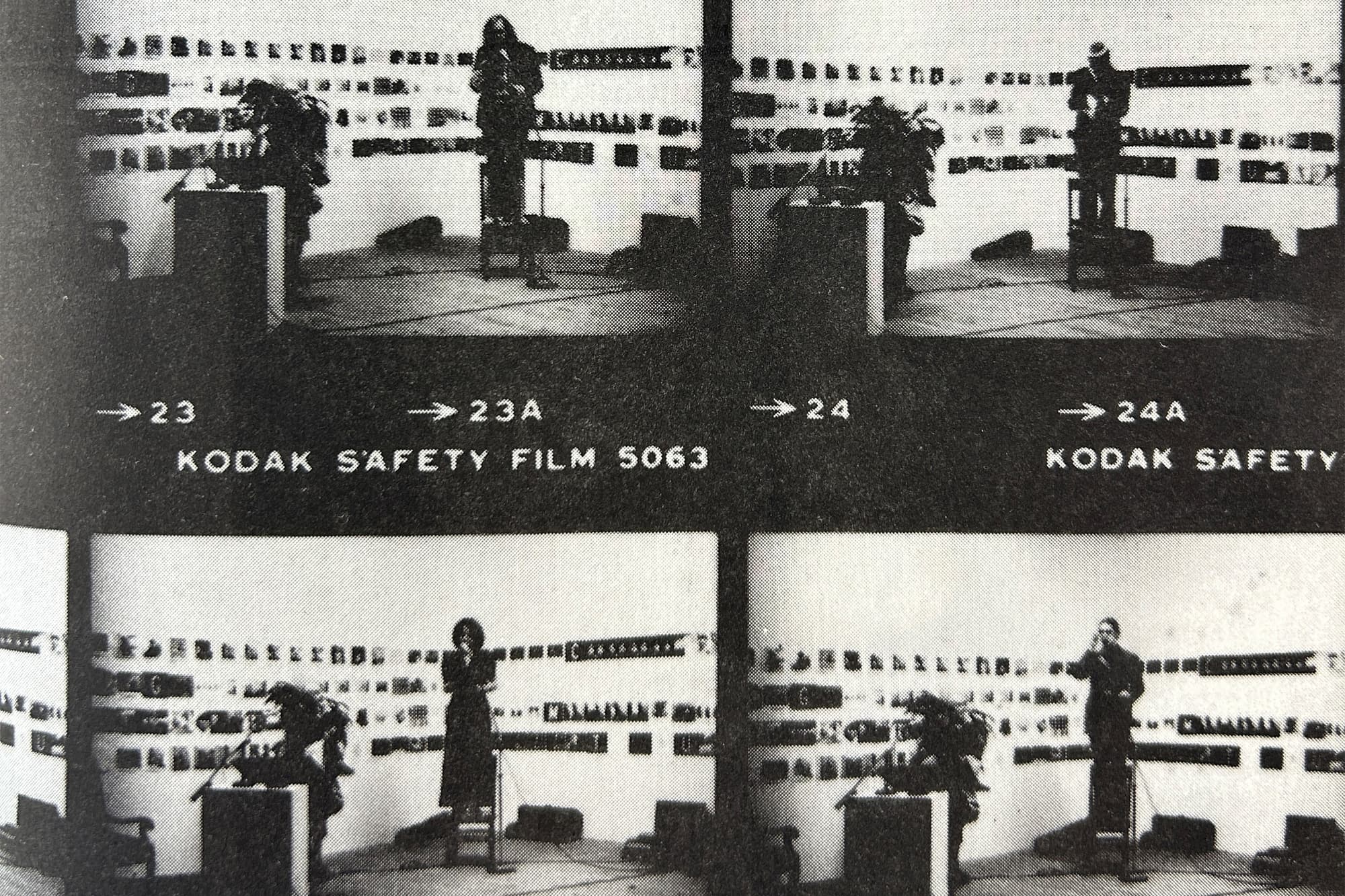 A black-and-white contact sheet showing four grainy photographs of a live performance. In each frame, a performer stands on a small stage behind a podium or table, with another figure and equipment nearby. The backdrop is covered with rows of abstract symbols, text, and graphic elements. Film markings, labels, and frame numbers, are visible around the sheet.