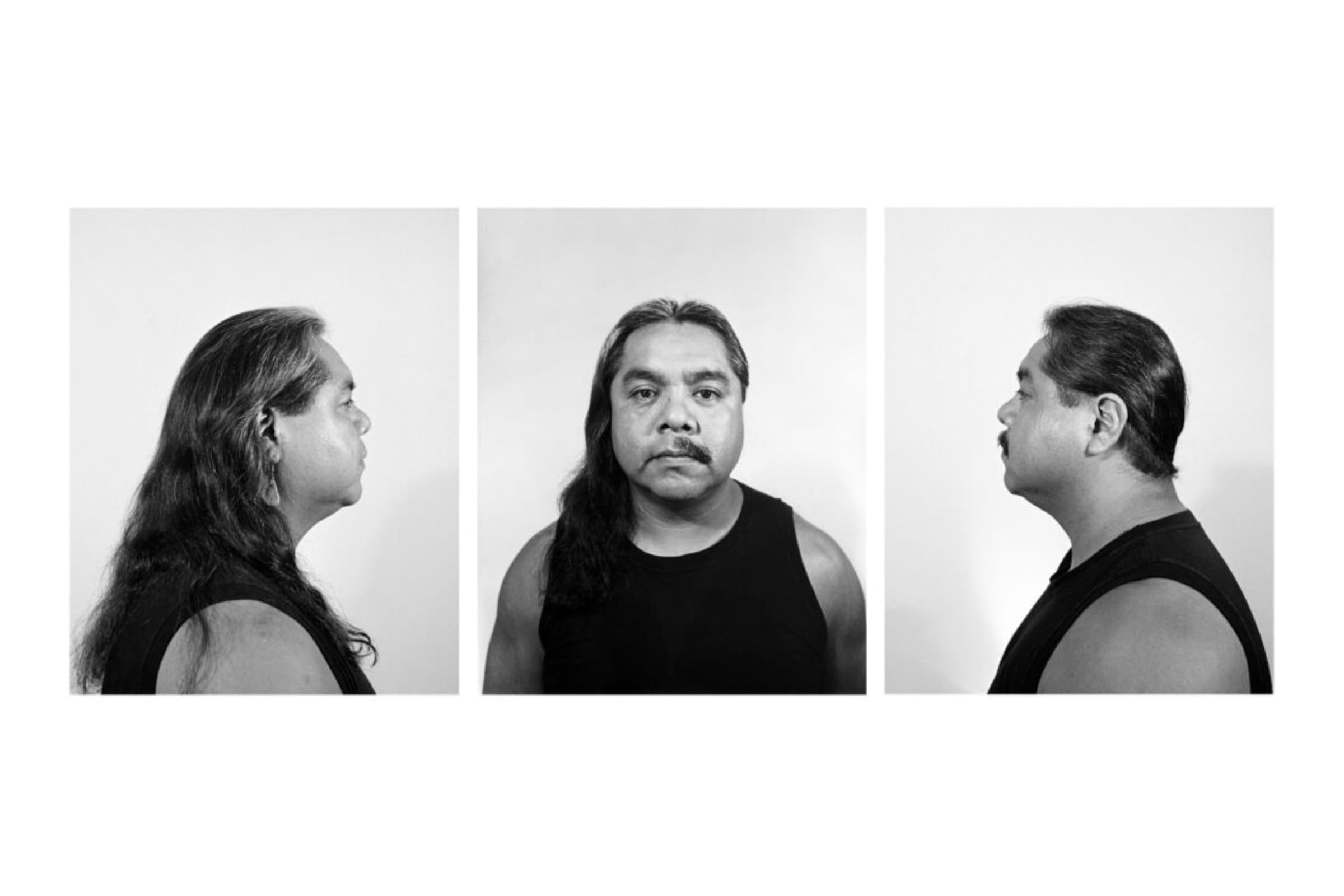 Three black-and-white photographic portraits of the same man are arranged side-by-side against a plain, light background, evoking the clinical style of a mugshot or an ethnographic study. In the left panel, the man is shown in a sharp profile facing right, displaying his long, dark hair flowing down his back and a distinct hanging earring. The center panel captures him facing directly forward with a neutral expression, his long hair parted down the middle with one side draped over his shoulder and half of his mustache is completely shaved. The right panel shows him in profile facing left, with his hair appearing tucked back or shorter to highlight his facial structure and mustache. Across all three frames, he is dressed in a simple, dark, sleeveless tank top.