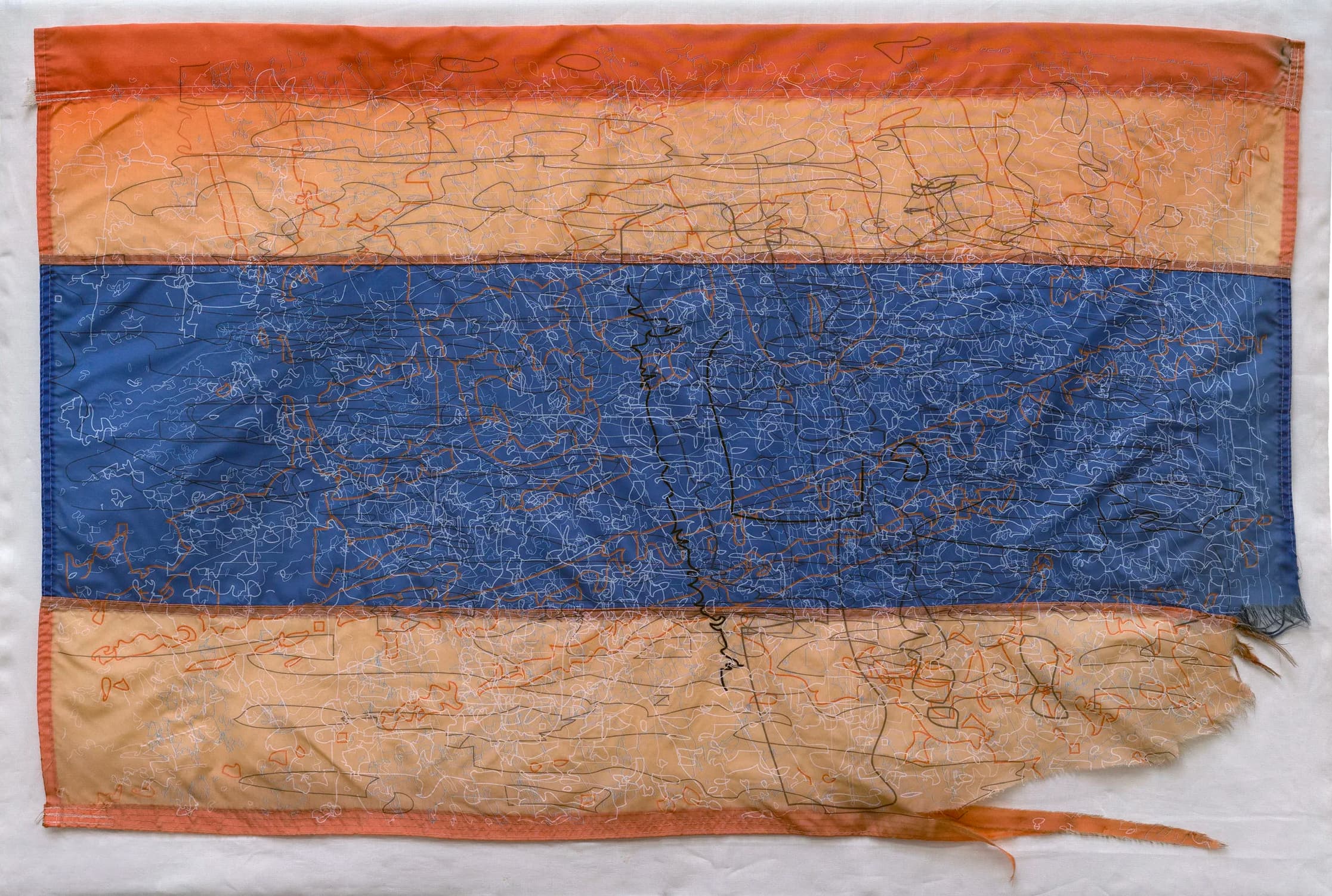 A water station flag featuring three horizontal stripes: orange on the top and bottom, and blue in the middle. The fabric is covered in asymmetrical blue, orange, and white threads sewn in all directions, and the lower right corner is frayed and torn.