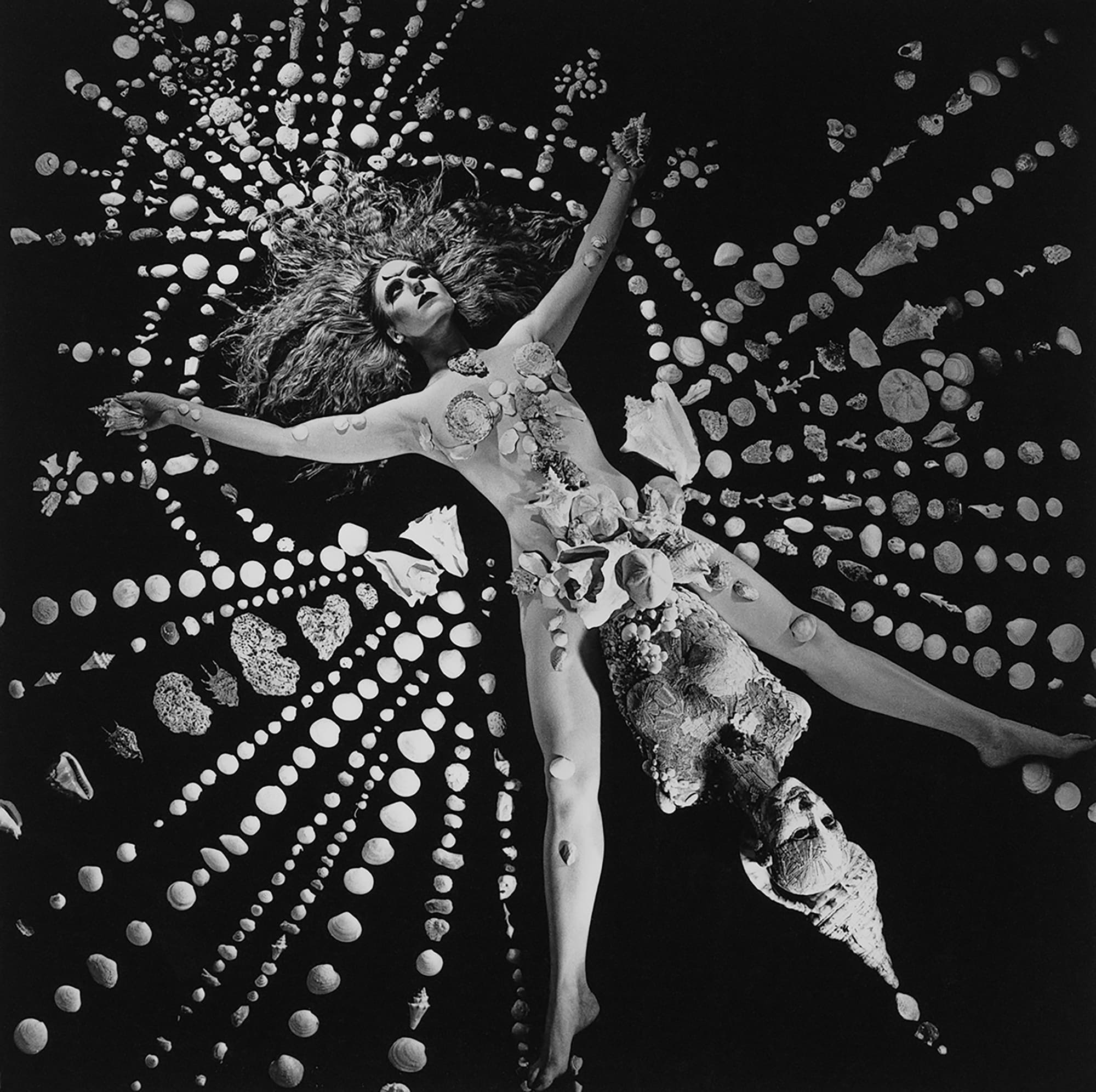 Black and white photograph of androgynous white person laid flat with arms and legs stretched in star orientation covered and surrounded by shells of varying size and texture