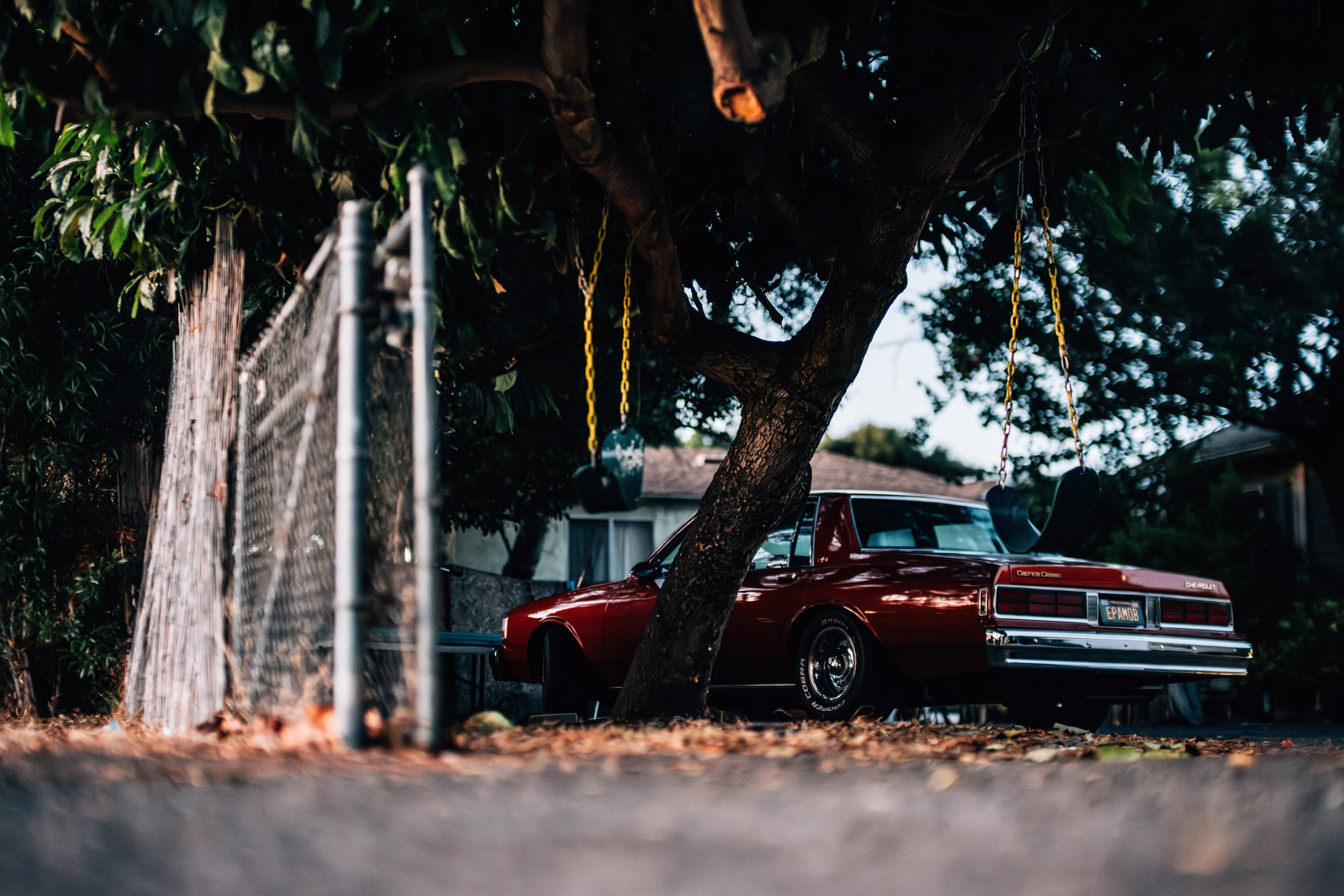 Image taken from the ground, with a grey metal fence on the left foreground and a cherry-red car filling the center and right side of the image. There are two tire swings suspended from a tree with yellow chain links.