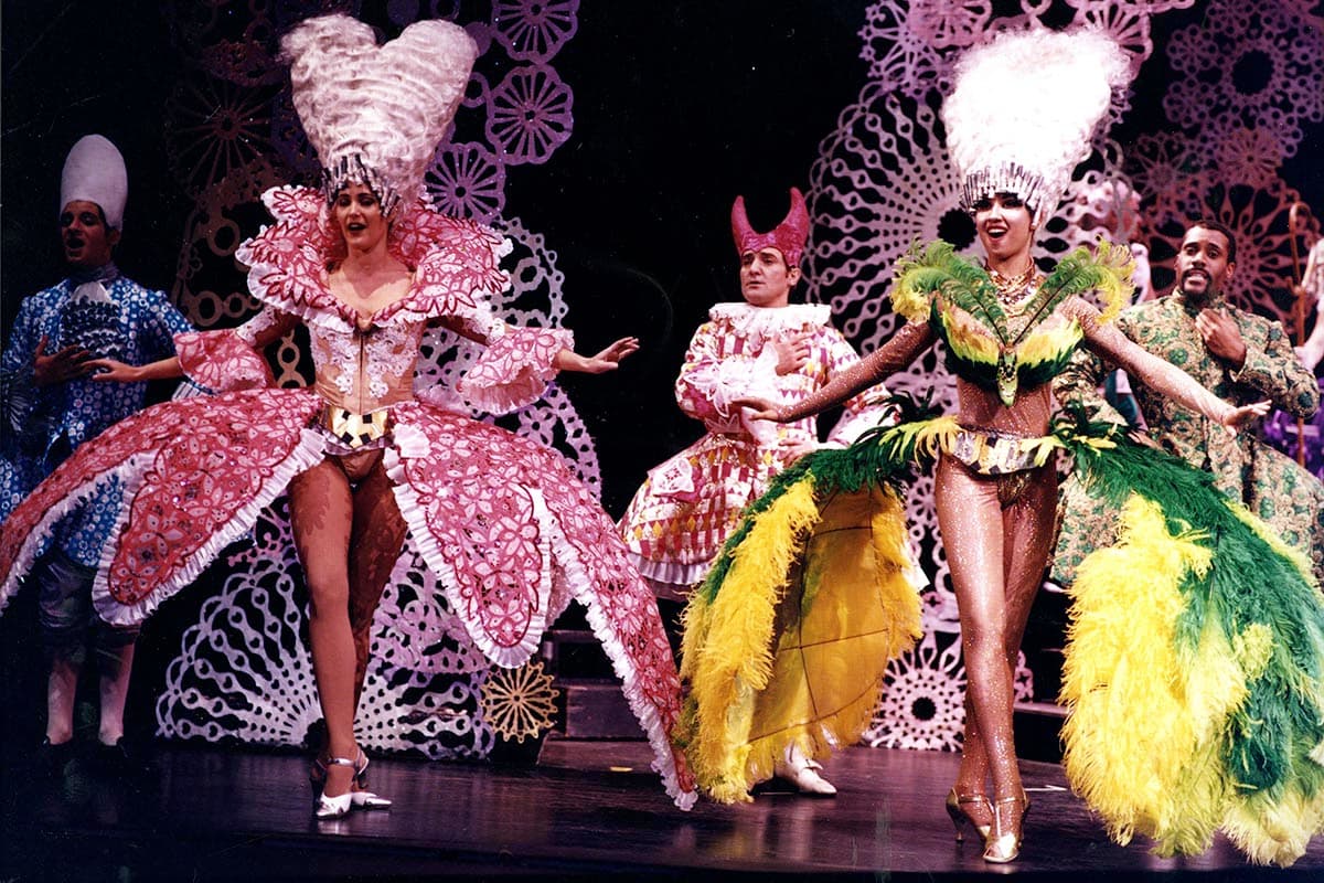 A group of performers posing on a dark stage in extravagant, colorful costumes. On the left, a performer wears an oversized pink lace-patterned hoop skirt and a towering white heart-shaped wig. To the right, another performer smiles while modeling an elaborate costume adorned with vibrant yellow and green feathers, topped with an oversized white wig. Other cast members in patterned outfits, including one wearing a red-horned headpiece, stand behind them against a background decorated with white lace-like patterns