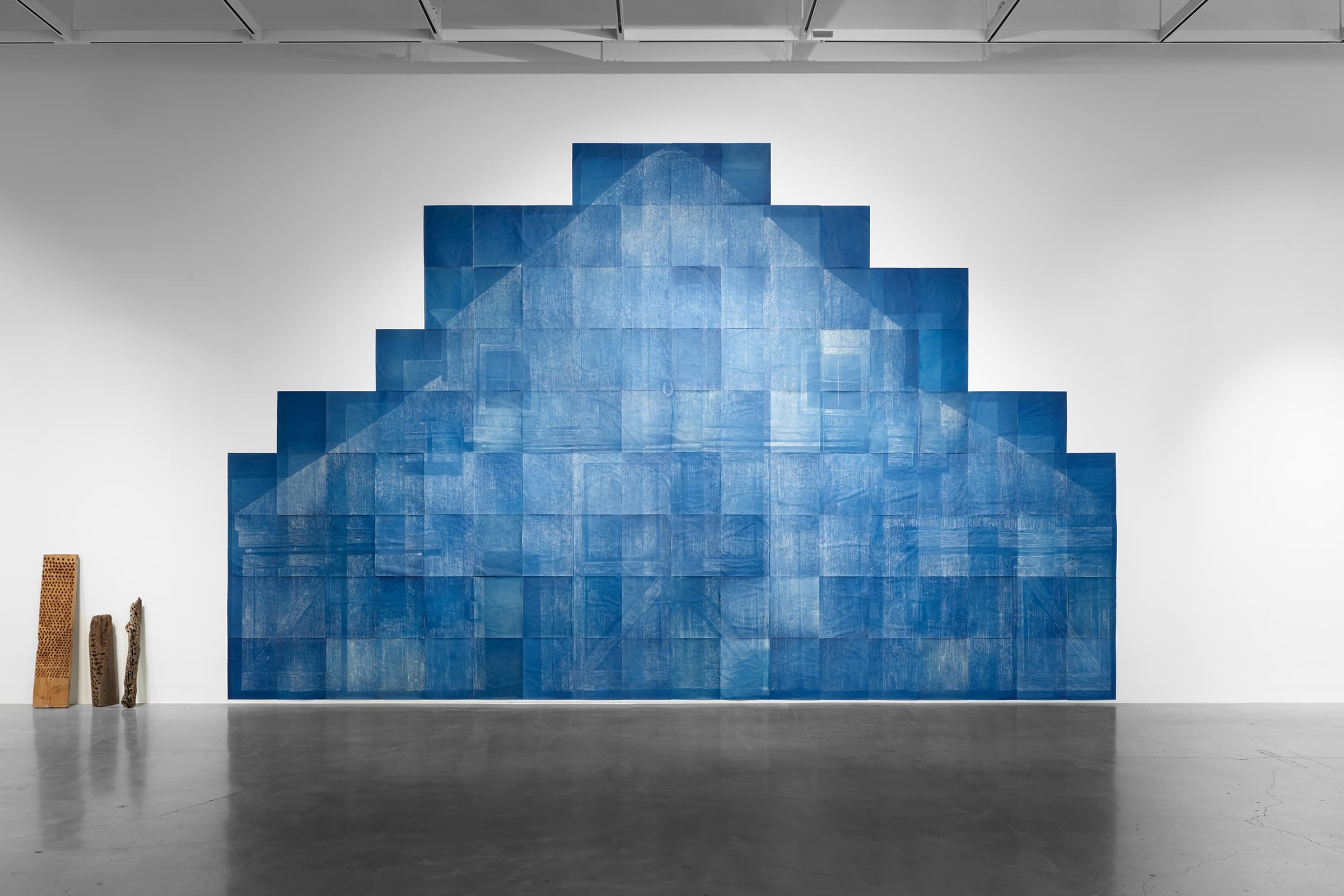 Installation view of a large, stepped blue geometric wall artwork composed of overlapping rectangular panels, with three narrow sculptural forms standing on the floor at left in a spacious gallery.
