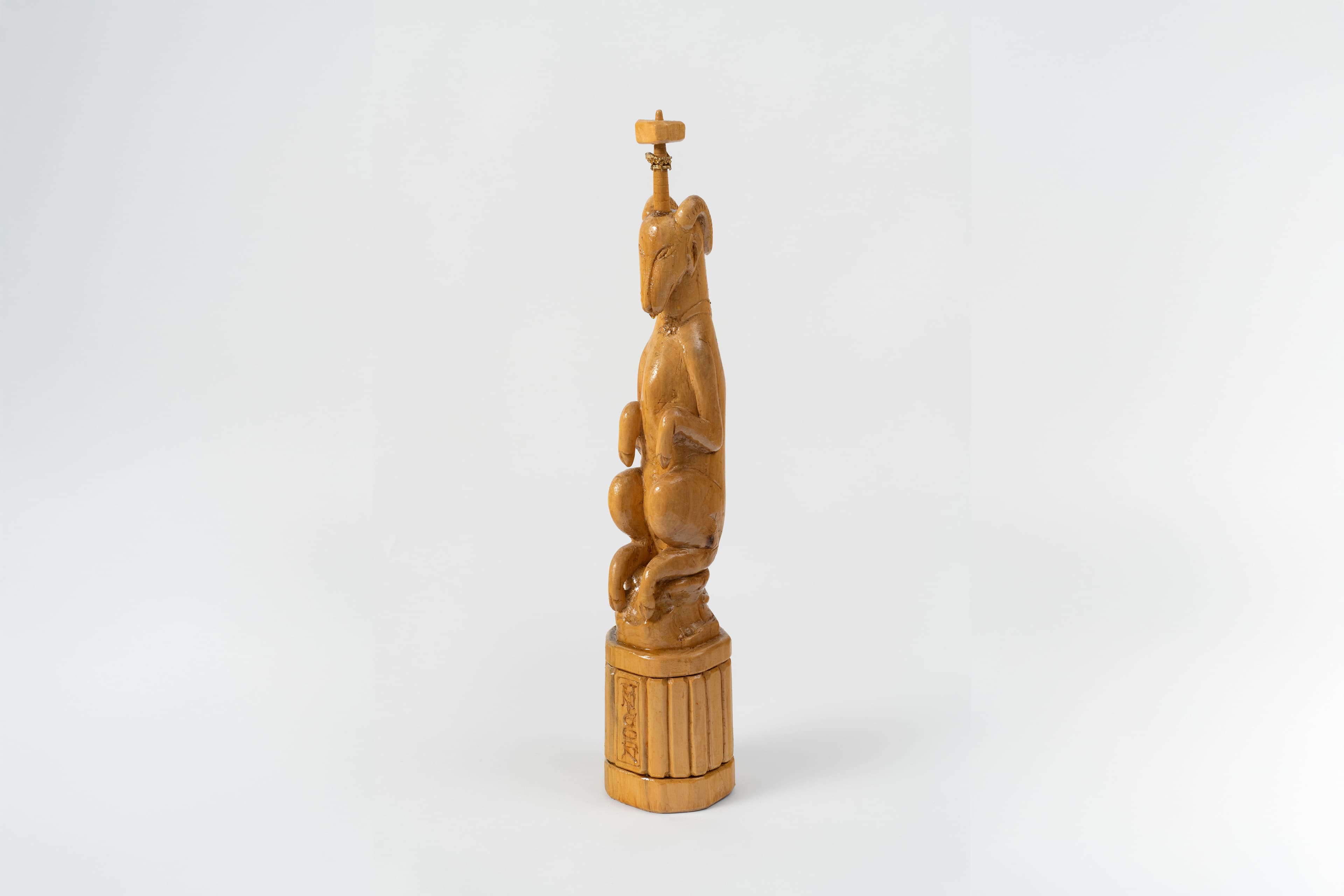 A tall, slender sculpture of a stylized animal, similar to a greyhound, perched on a decorative cylindrical base. The light-colored wood carving depicts a creature with long, smooth lines, rabbit or goat-like ears and paws, and a tapered snout, sitting upright in a crouching posture. Atop its head stands a thin, vertical rod capped with a small, cross-like horizontal element. The entire figure rests on a fluted, columns-style circular pedestal that features vertically carved panel lines and a faint rectangular inscription carved into one side.