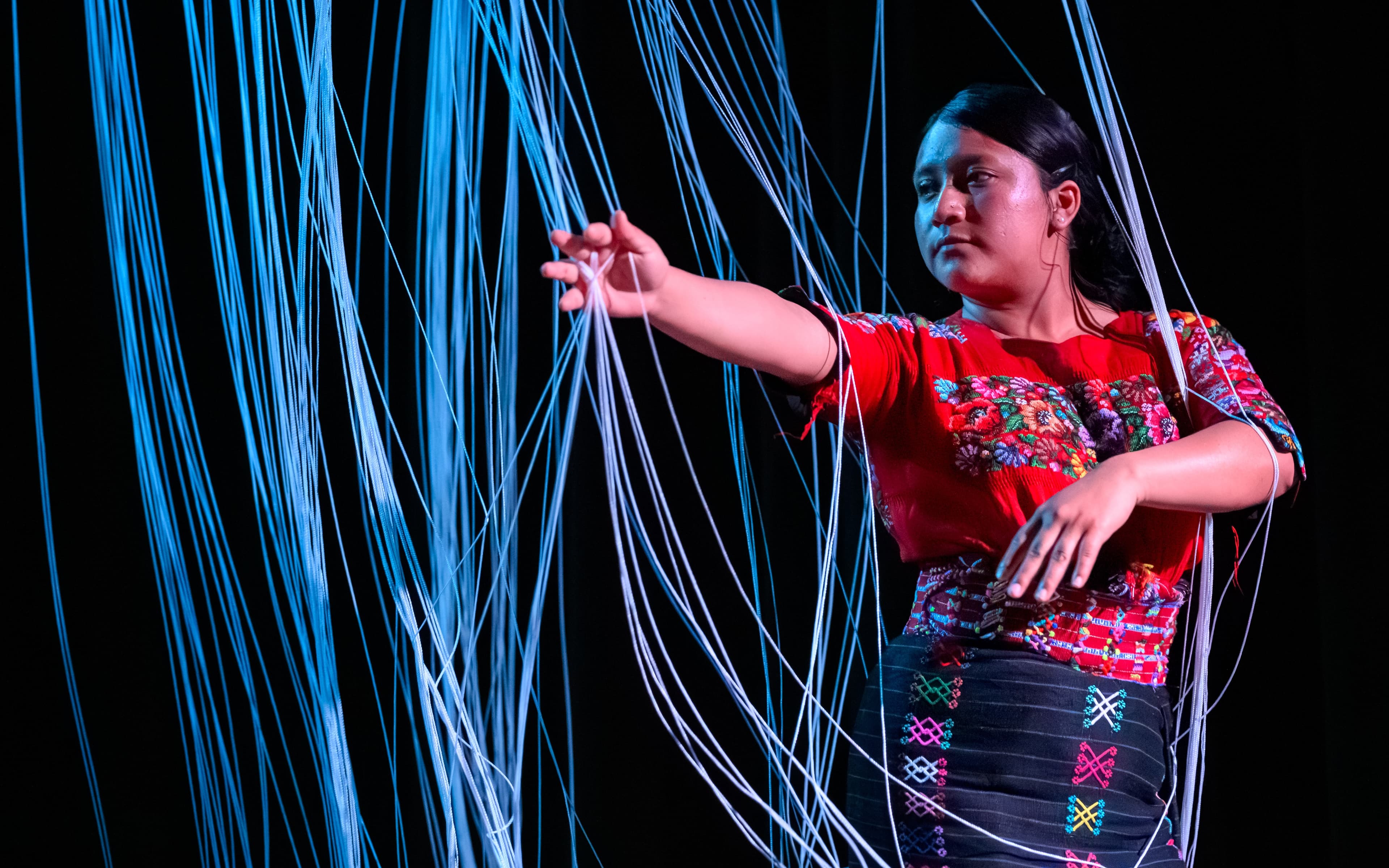 A young Maya Mam woman is dancing in a curtain of white strings. She is wearing a red huipil with brightly colored flowers across the chest, a red belt and black skirt adorned in colorful designs: pink, green, blue, white, orange. She is looking to her right and her right arm is outstretched.