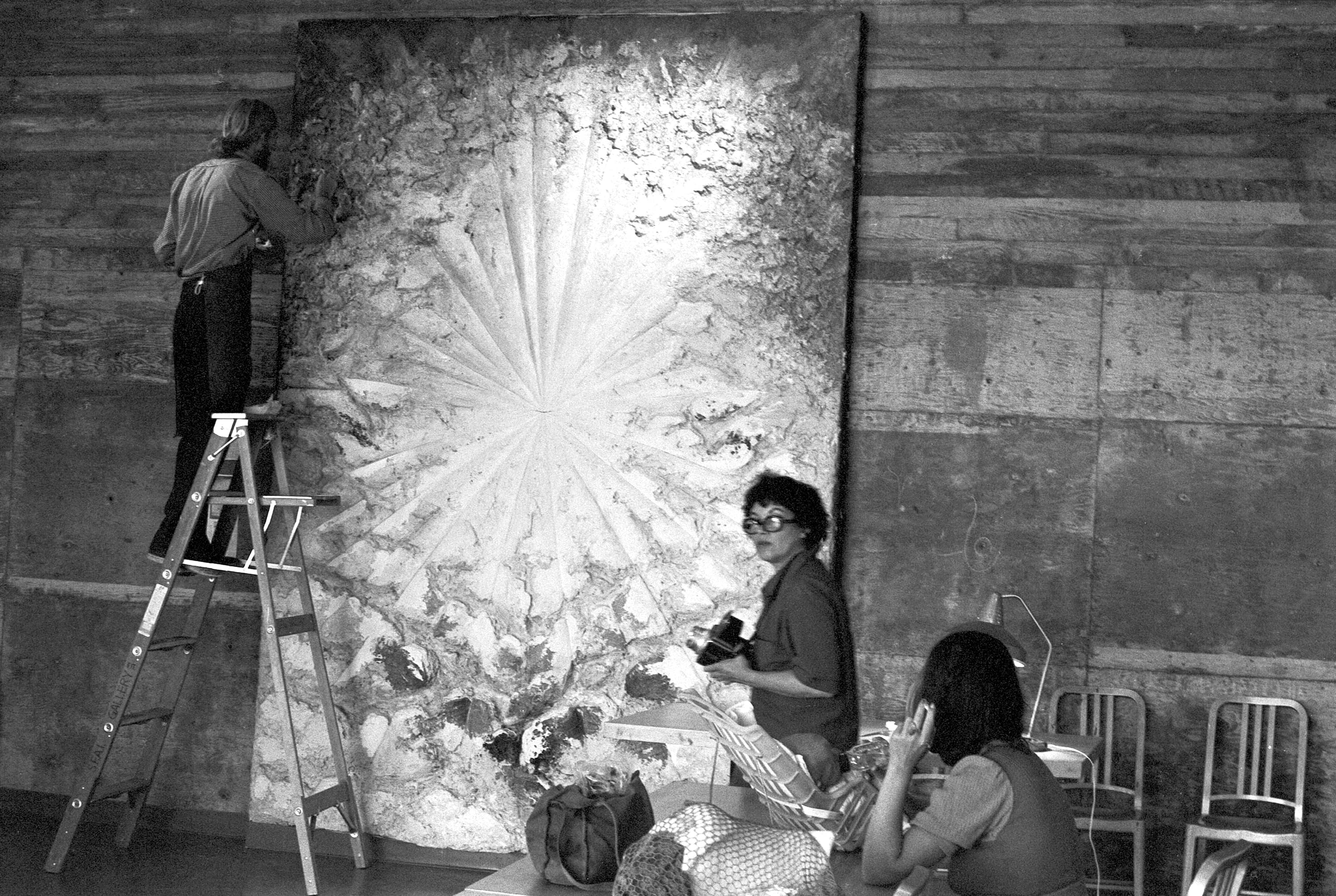 A large-scale, abstract, heavily impasto painting hangs on a concrete wall, the artist stands to its right.