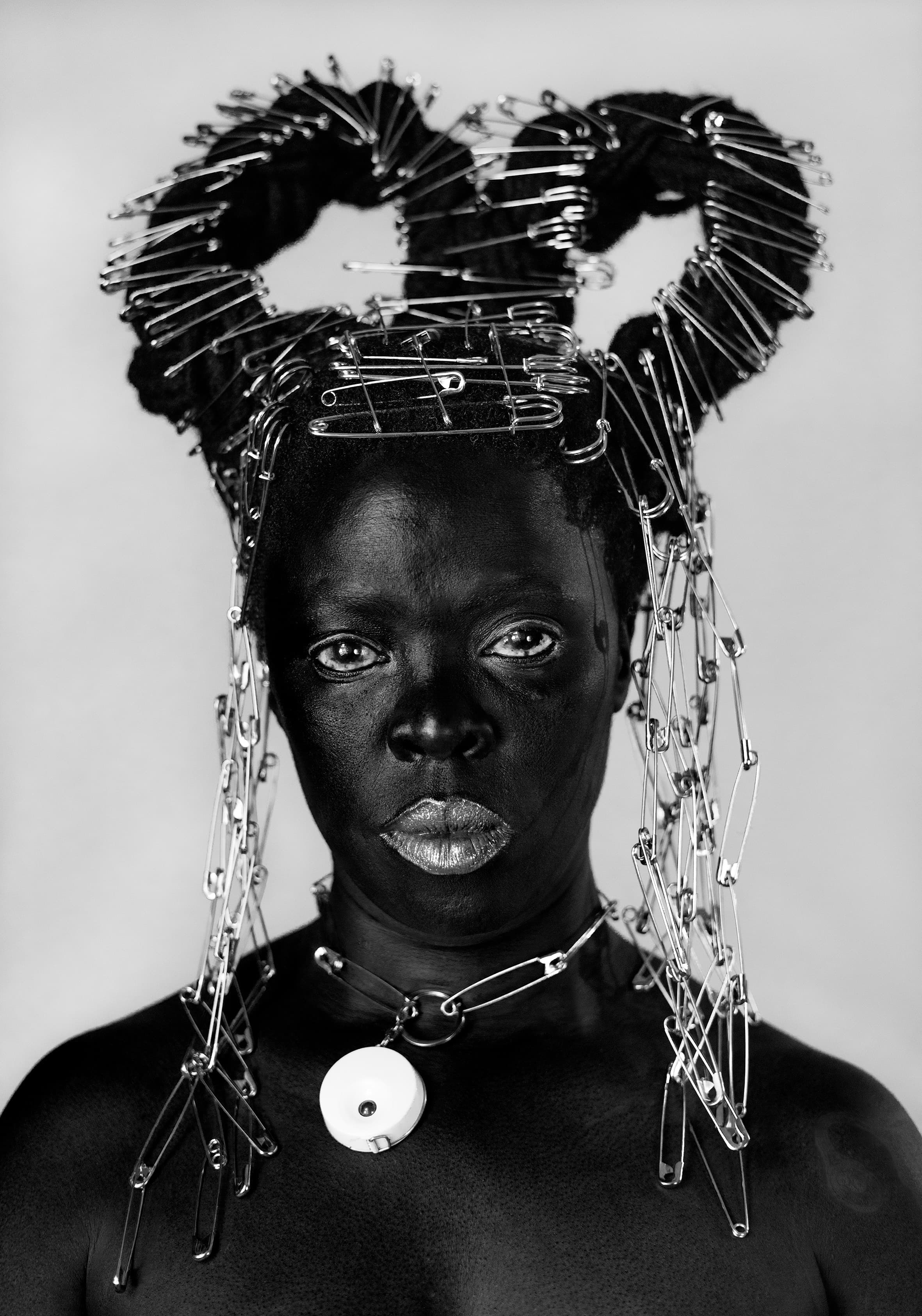 Self portrait of photographer Zanele Muholi with a necklace and headpiece made of safety pins wrapped around their locked hair and draped down the sides of their face.