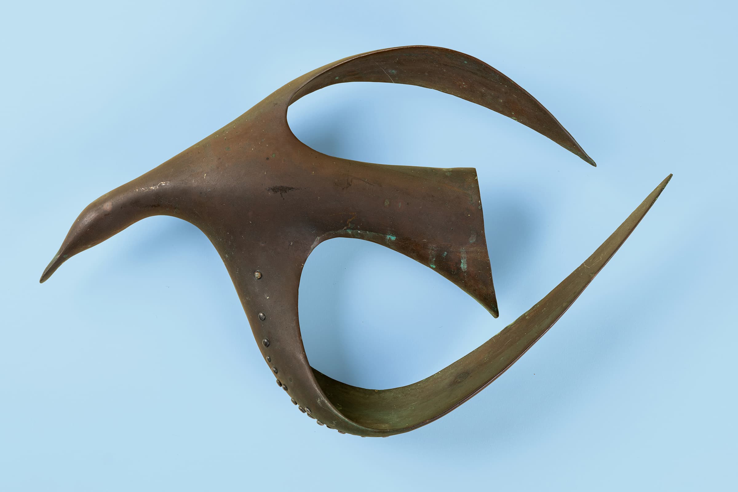 Abstract, biomorphic bronze sculpture of a stylized bird in flight. The sculpture features a smooth, patinated brown surface with subtle green verdigris highlights. Its fluid form is defined by swooping, dynamic lines and elegant negative spaces that form the wings and tail. Small, raised rivets line the lower curved section of the front wing, adding a textured, structural element to the otherwise sleek, sweeping design.