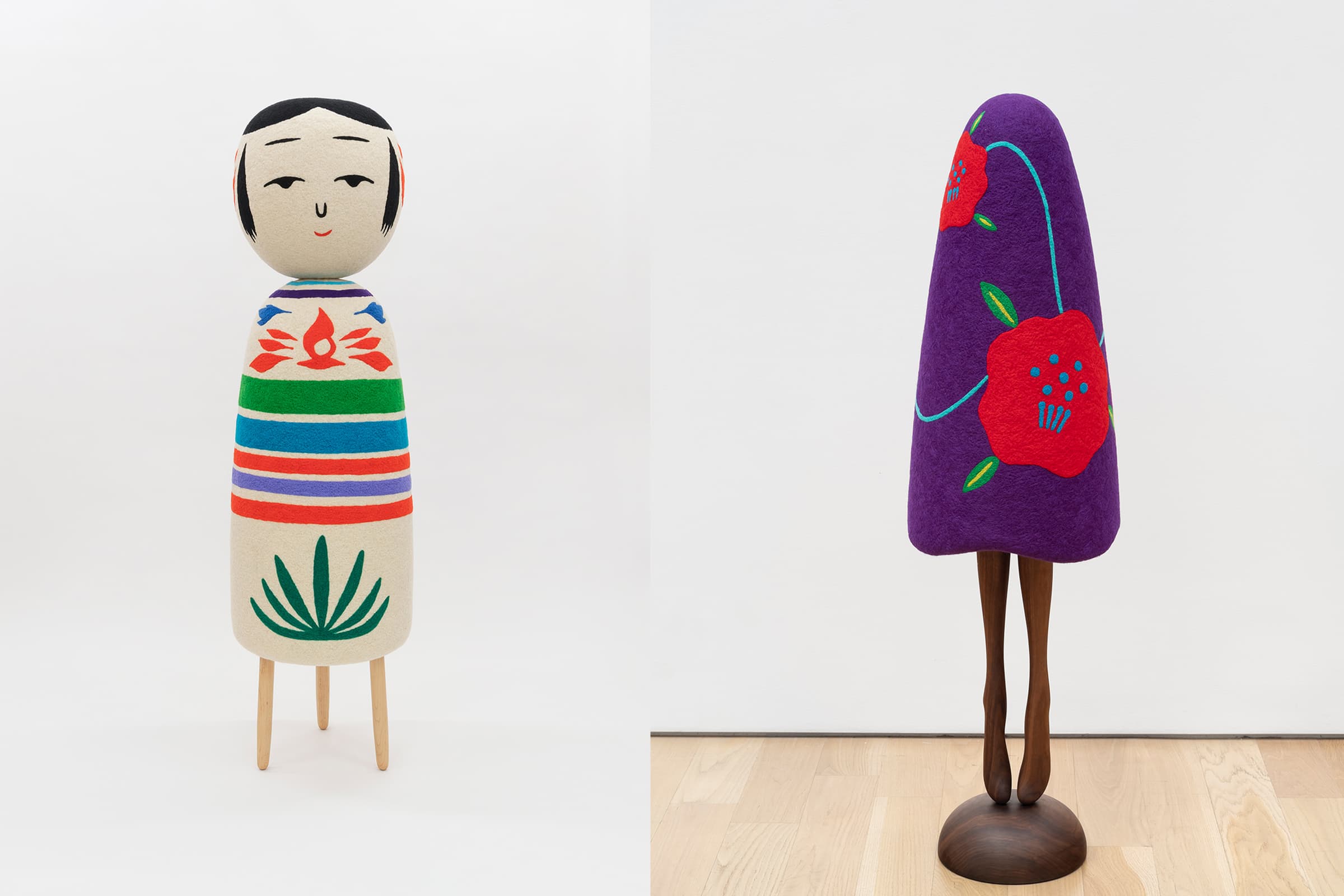 To the left: A stylized, cylindrical sculpture resembling a traditional Japanese kokeshi doll, featuring a friendly face, vibrant horizontal stripes in her body, and a green palm emblem at the bottom of her body before wooden legs. To the right: A sculpture featuring a slender wooden base resembling humanoid legs, topped with a tall, rounded purple cone embroidered with large red flowers and green leaves connected via a thin stitched blue thread.