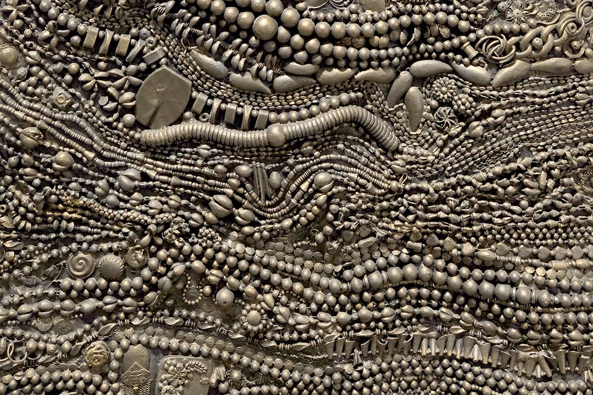 Detail of a sculptural dress-like form covered in intricate layers of beads, chains, medallions, and metal ornaments.