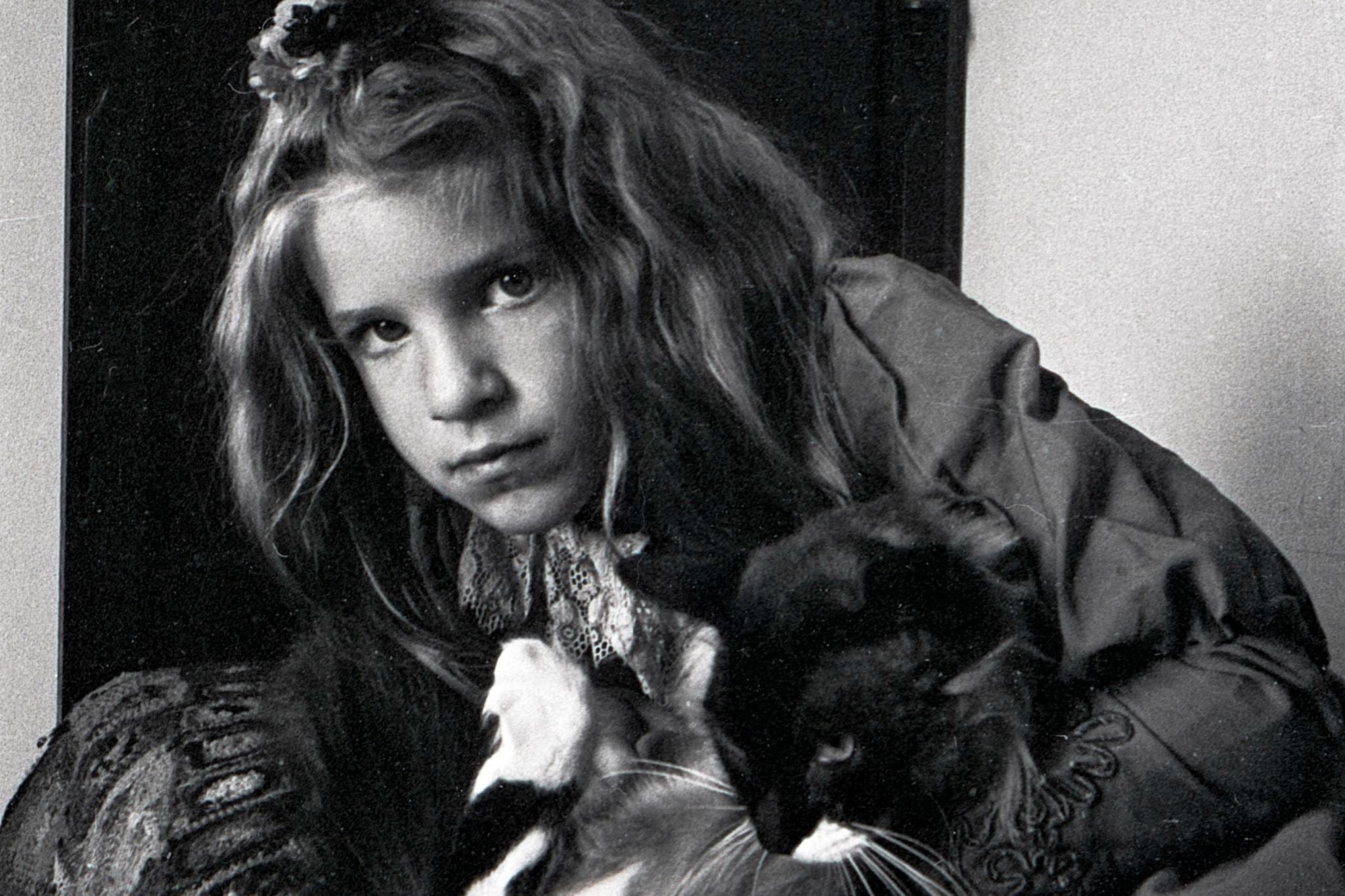 Girl with cat. A close-up black and white portrait of a young girl with long, wavy hair and a serious expression. She is looking directly into the camera while holding a fluffy black-and-white cat close to her chest. She wears a dress with an intricate lace collar detail.