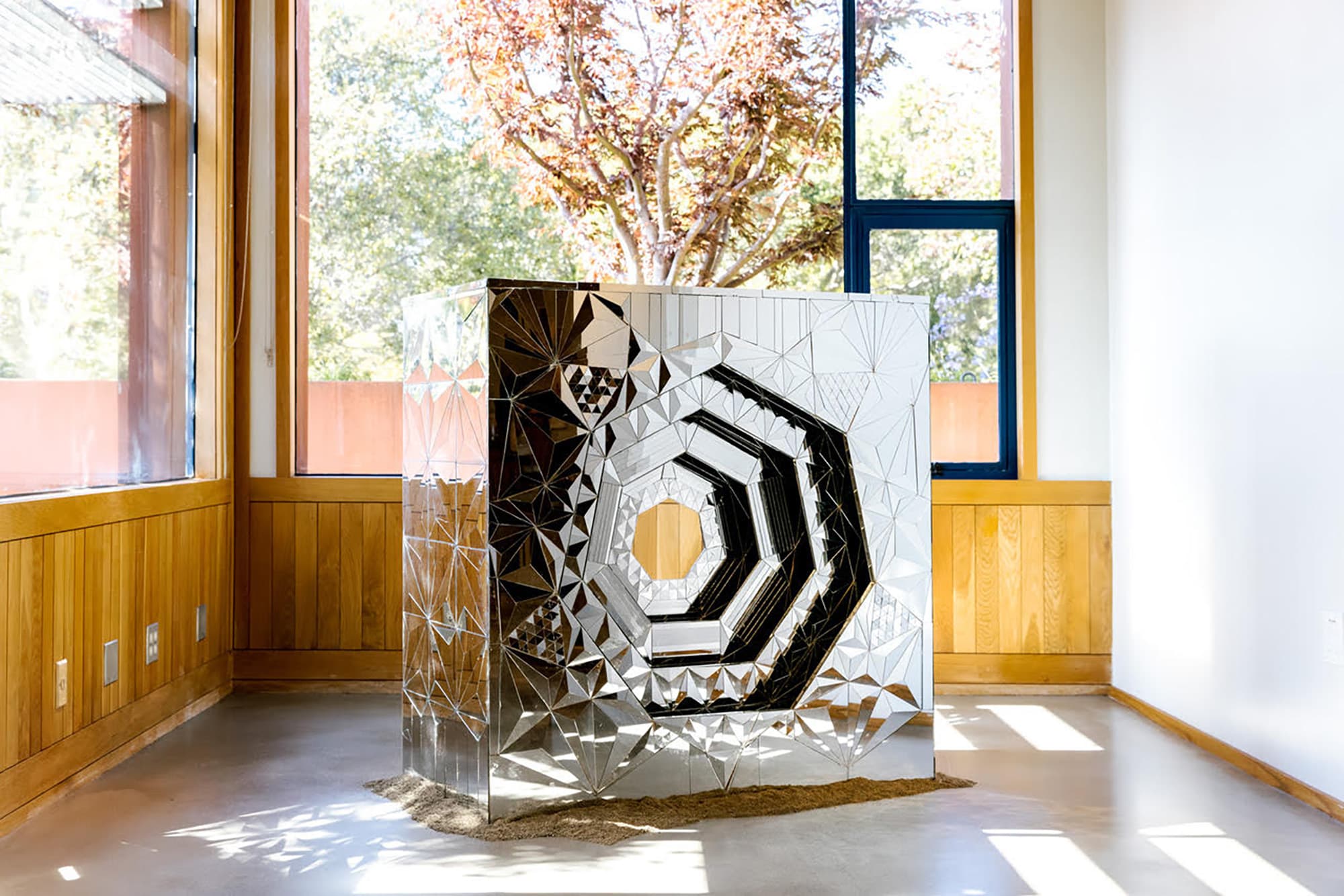 A large mirrored geometric sculpture with a faceted, kaleidoscopic surface sits in a sunlit room. Its angular paneling forms a deep, concentric tunnel opening in the center.